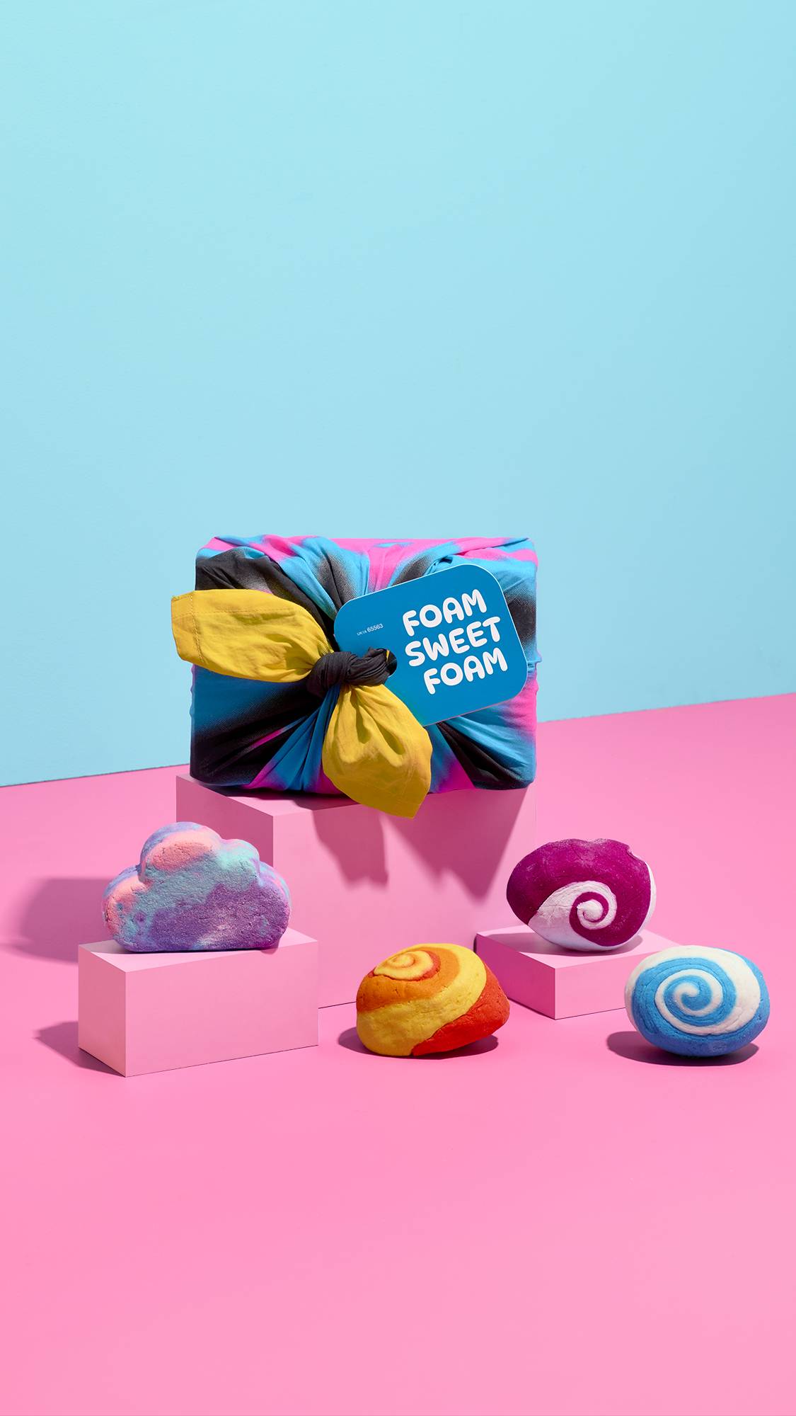 A fabric-wrapped "FOAM SWEET FOAM" gift set sits on a central block surrounded by four colourful bubble bars—a pastel cloud shape and swirled blue, orange, and purple bars—arranged on pink pedestals against a blue backdrop.
