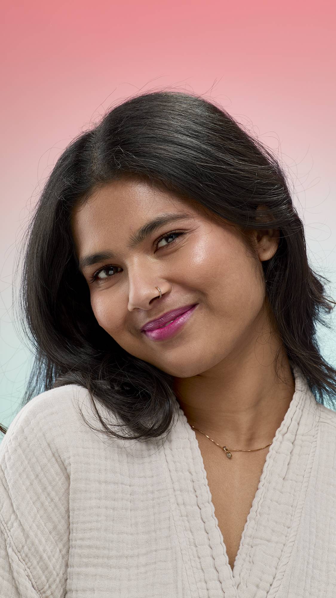 A person with long dark hair and a nose ring smiles at the camera, wearing the shimmering, deep berry-purple Frankenstain lip stain. They are wearing a white textured robe against a soft pink-to-blue gradient background.