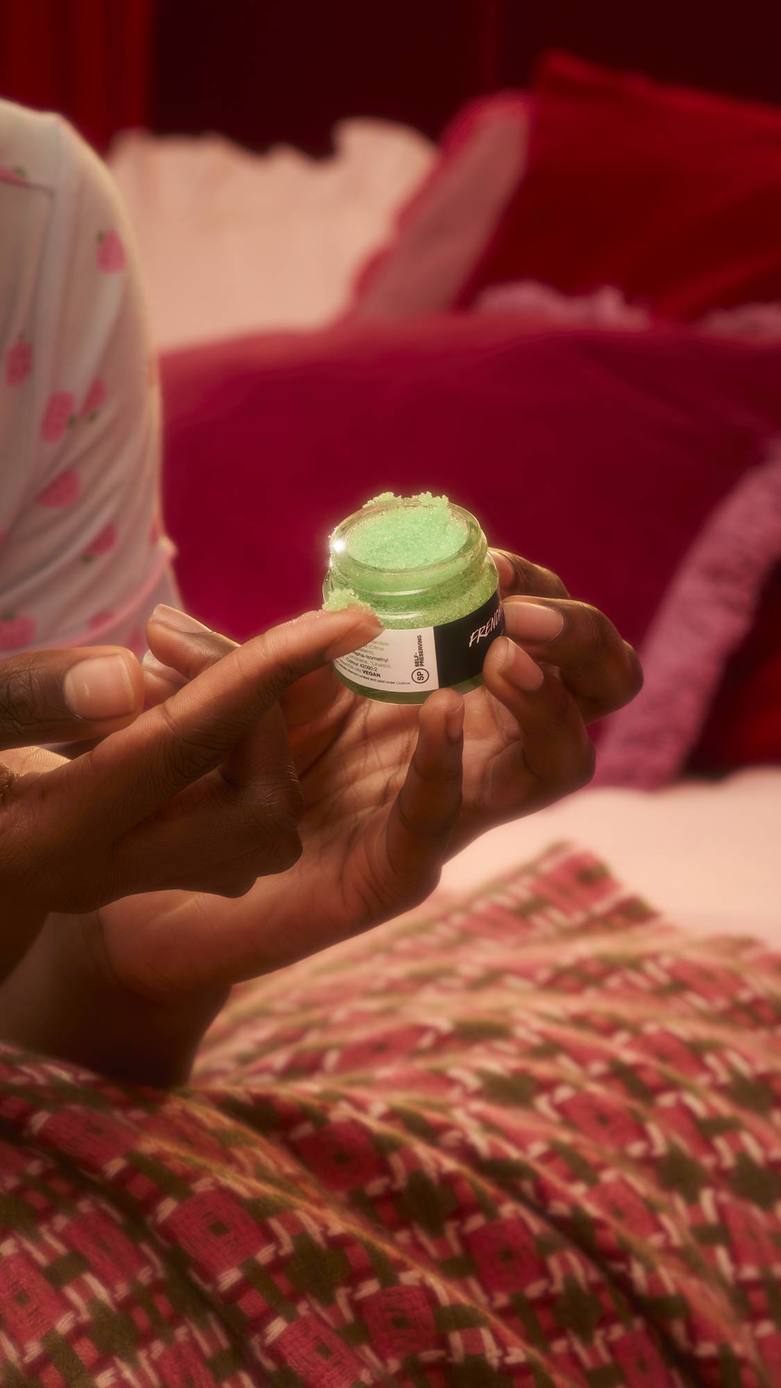 A woman in pink strawberry pyjamas holds an open jar of green "French Kiss" lip scrub while reclining on a bed. She smiles as she prepares to apply the product with her finger in a cosy, candlelit bedroom setting.