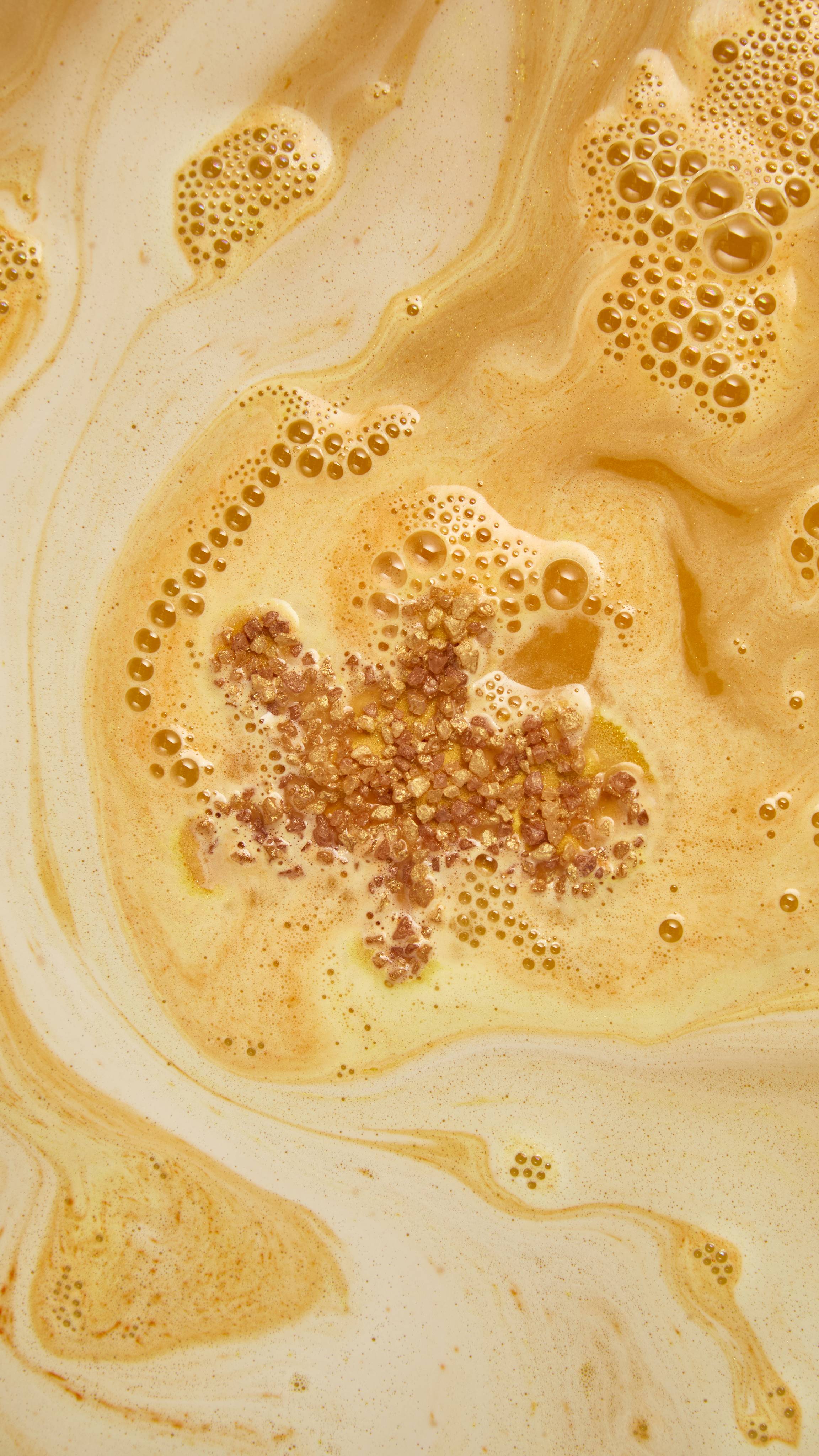 Golden-brown crystals from a gingerbread man bath bomb float at the centre of rich, swirling caramel and cream-coloured foam, surrounded by fine bubbles in warm amber bathwater.