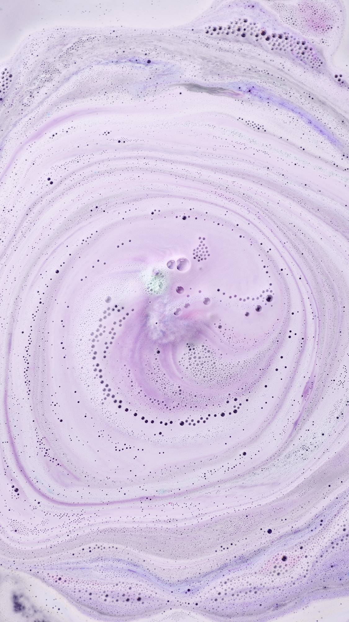 
An aerial view of pale lilac bathwater covered in swirling patterns of white and purple foam. Tiny bubbles and darker purple droplets create intricate trails across the surface, spiralling towards the fizzing "Goddess" bath bomb.