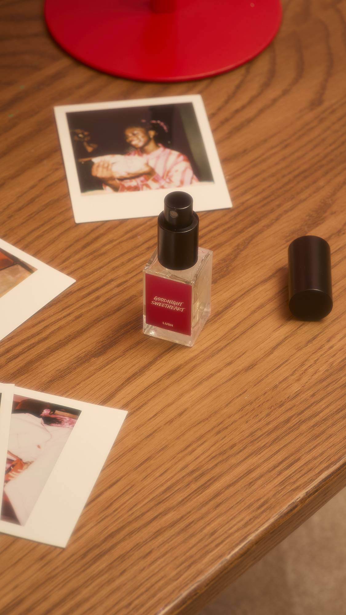 A bottle of Lush "Goodnight Sweetheart" pillow spray perfume, with its cap removed, sits on a wooden surface alongside several Polaroid photographs. The clear glass bottle features a vibrant magenta label with white text.