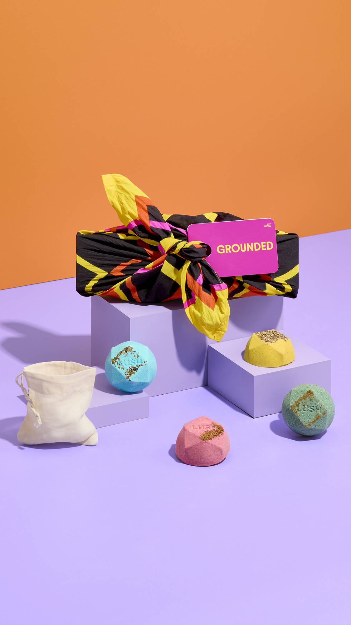 A knot-wrapped gift tagged "GROUNDED" sits on lilac plinths against an orange background, surrounded by four colourful Lush bath bombs in blue, yellow, green, and pink, alongside a small beige cotton pouch.