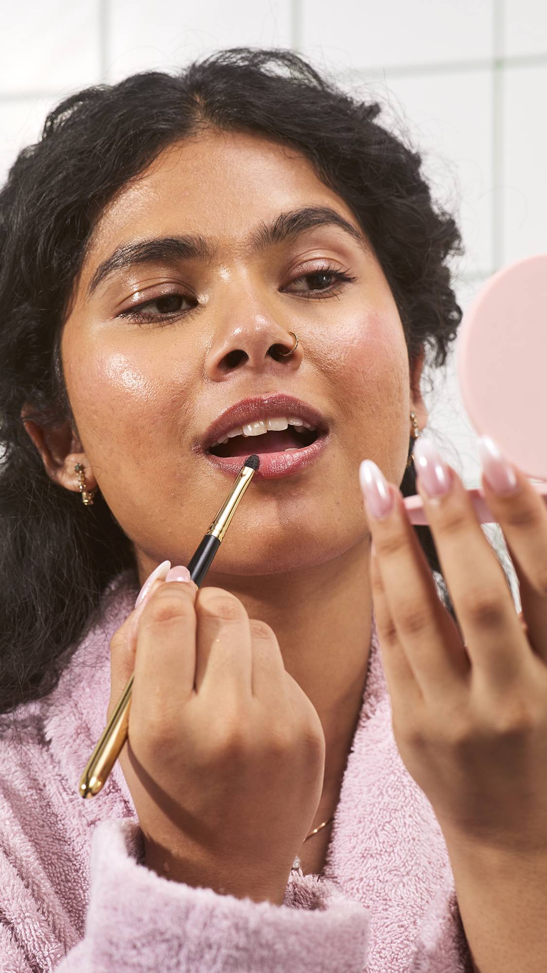 A model wearing a pink fluffy bathrobe holds a pink compact mirror and uses a small makeup brush to apply Lush "Hot Chocolate Orange" jelly lip mask to their lips, set against a white tiled bathroom wall background.
