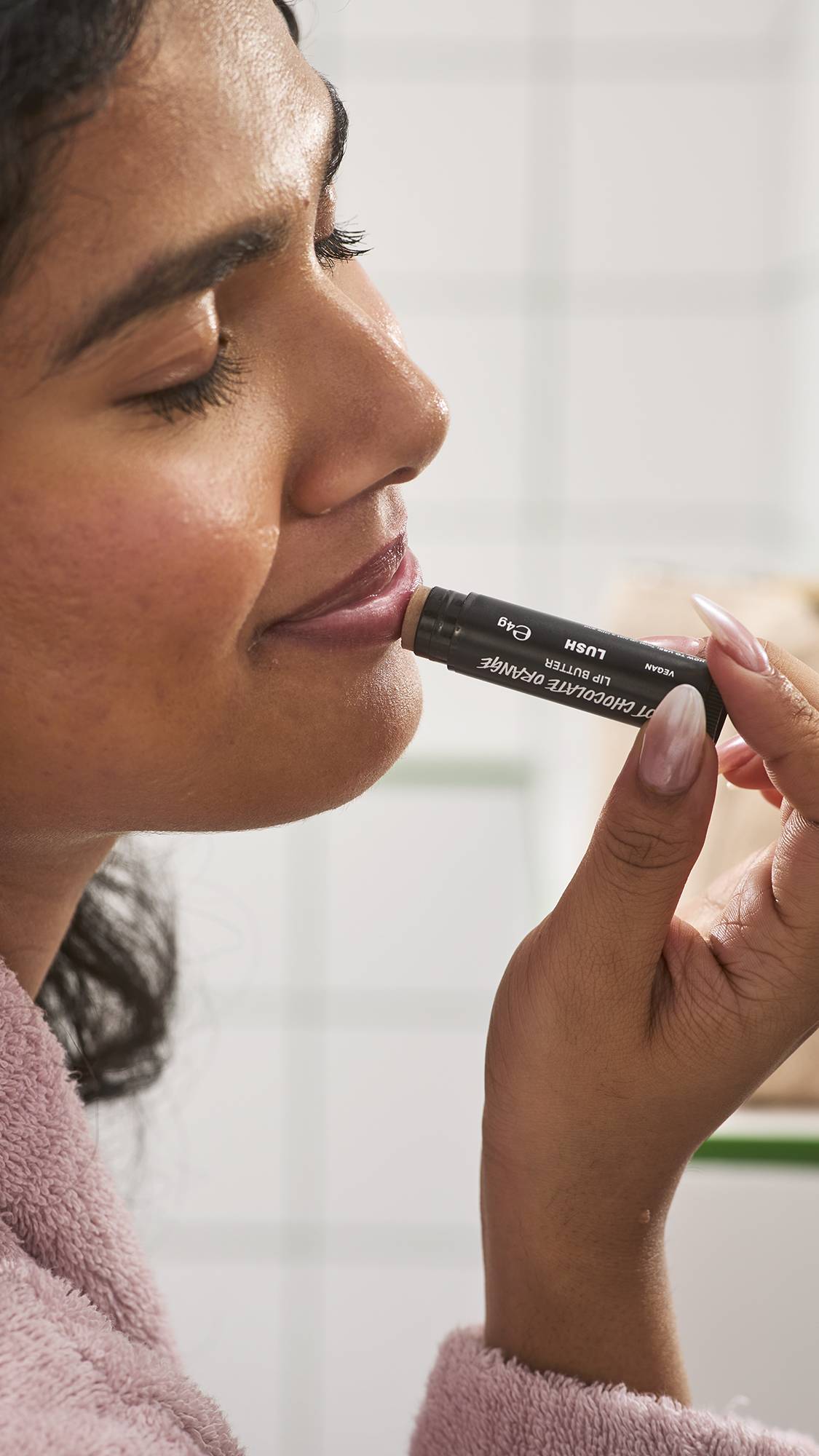 A side-profile close-up of a model in a pink bathrobe applying Lush Hot Chocolate Orange lip butter directly to their lips, set against a white tiled bathroom wall background.