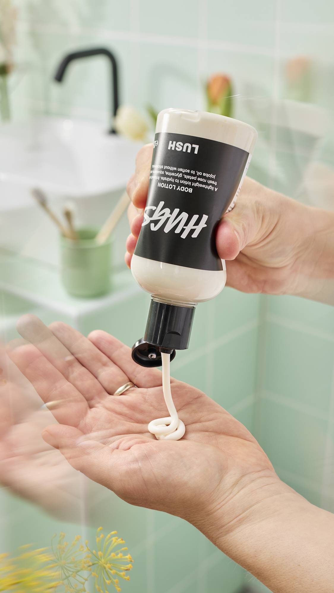 A person's hands squeeze a bottle of Lush "Hugs" Body Lotion, dispensing a swirl of thick white cream into their palm. The bathroom setting features pale green tiles, a blurred sink, and fresh flowers, suggesting a natural skincare routine.