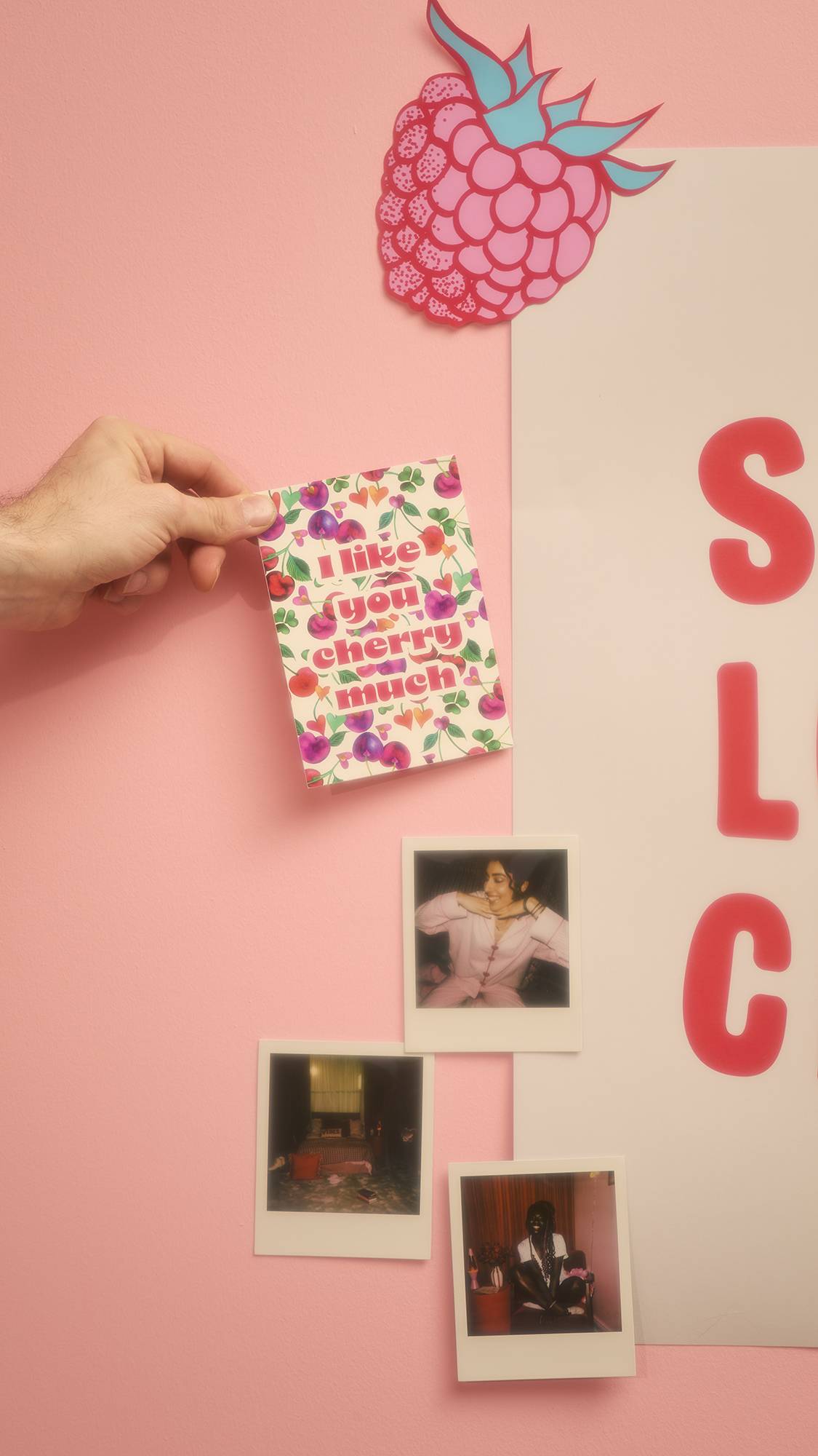 A hand holds the "I Like You Cherry Much" card against a pink wall decorated with a large raspberry cutout and Polaroid photos. Partial red text is visible on a poster to the right.