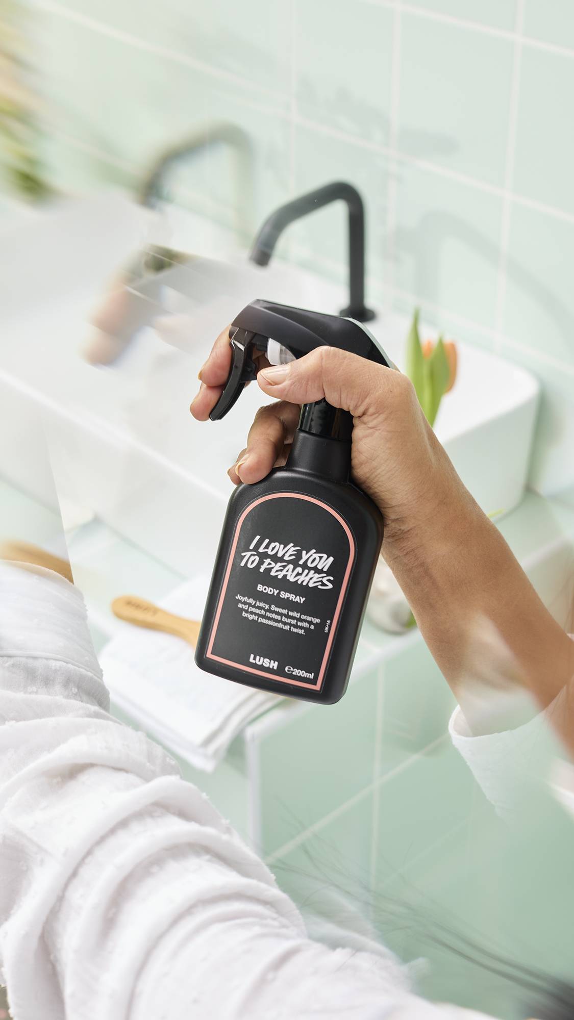 A model's hand holds a black bottle of Lush "I Love You To Peaches" Body Spray in a brightly lit bathroom. The black bottle has a peach-coloured border on the label, and the background shows a blurred white sink and pale green tiles.