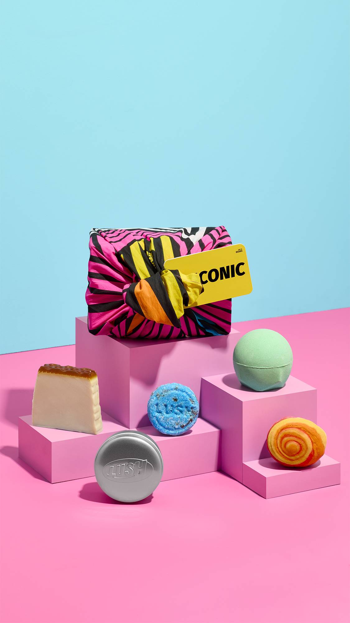 A pink and black striped "ICONIC" fabric-wrapped gift set sits on pink block pedestals alongside bath and body products, including a soap slice, blue shampoo bar, round silver Lush tin, mint green bath bomb, and red swirl bubble bar.