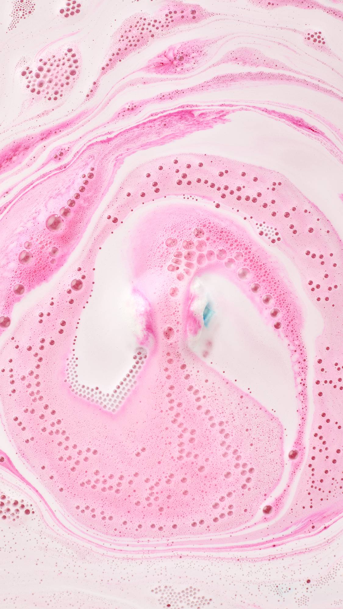 The Inclusion bath bomb dissolves in the bathwater. The pink, orange, and yellow geometric product releases thick, vibrant ripples of velvety foam across the water's surface.