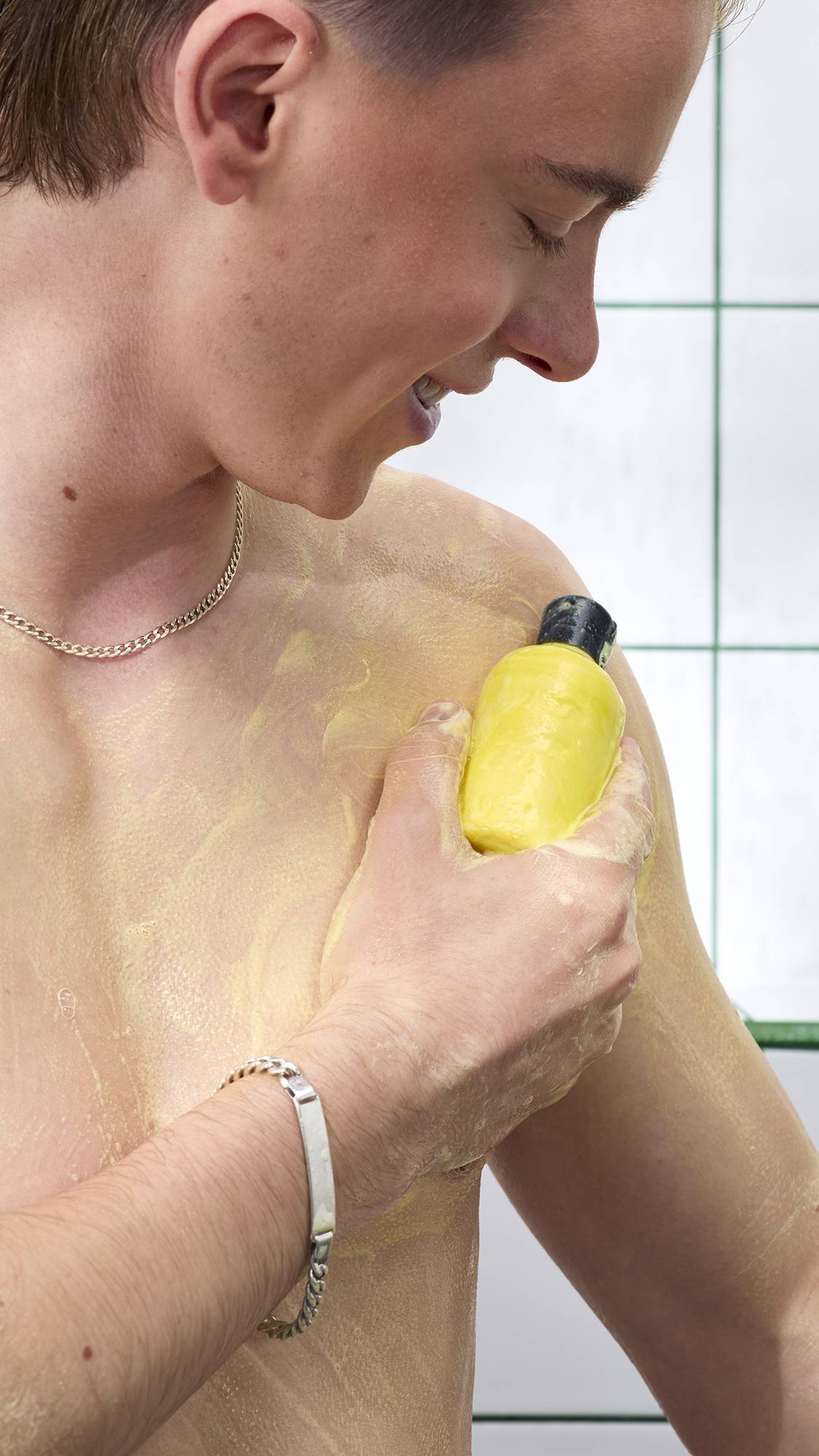 A side-profile shot of a smiling person lathering their shoulder in the shower with a yellow, bottle-shaped solid shower gel. Their chest is covered in yellow lather against a background of white tiles with green grouting.
