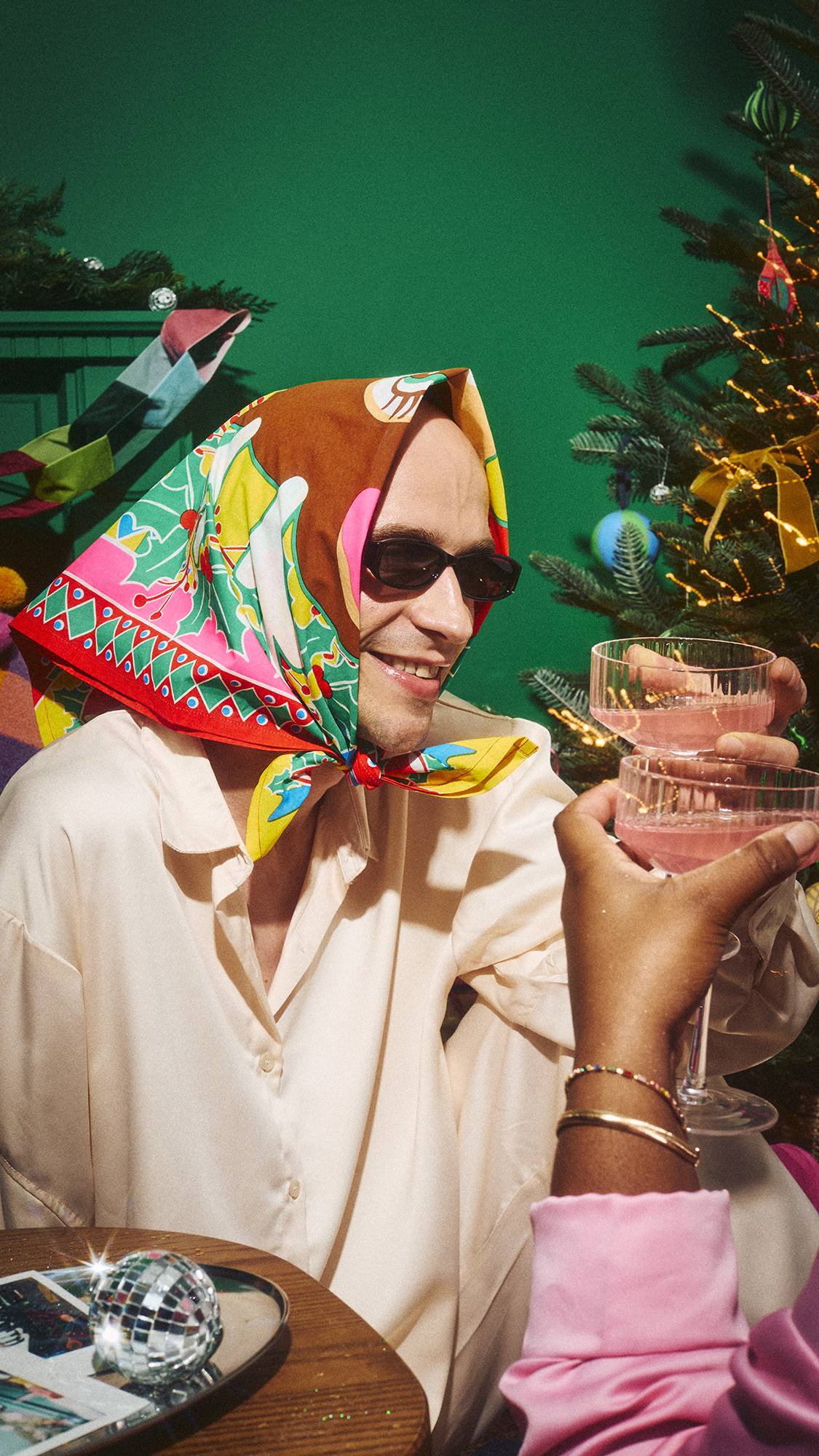 A smiling person wearing sunglasses and a colourful reindeer-patterned Lush fabric Knot Wrap, fashioned into a headscarf, toasts pink drinks with a friend in a festive room setting near a decorated Christmas tree.