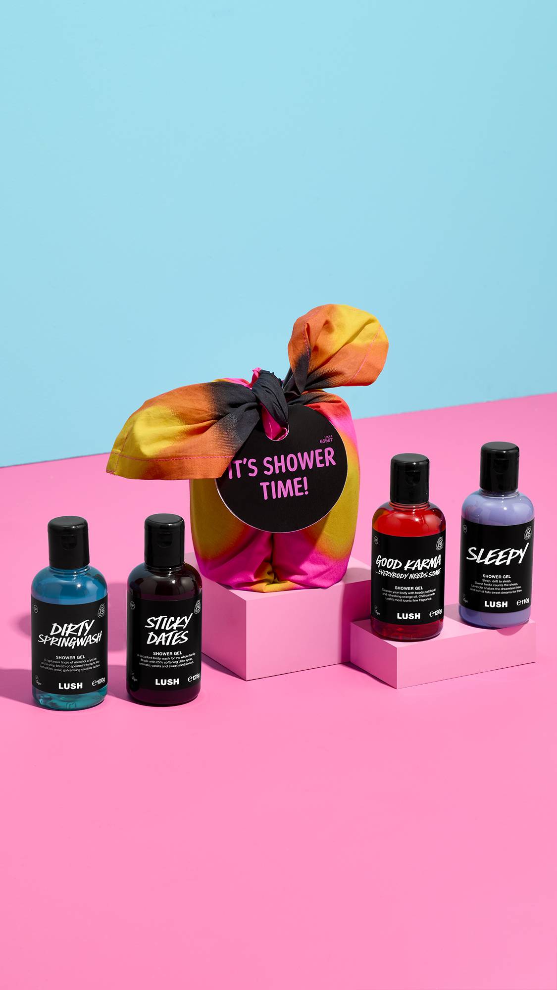 A neon tie-dye knot-wrapped gift tagged "IT'S SHOWER TIME!" sits on a pink plinth, surrounded by four Lush shower gels: Dirty Springwash, Sticky Dates, Good Karma, and Sleepy, set against a pink and blue background.