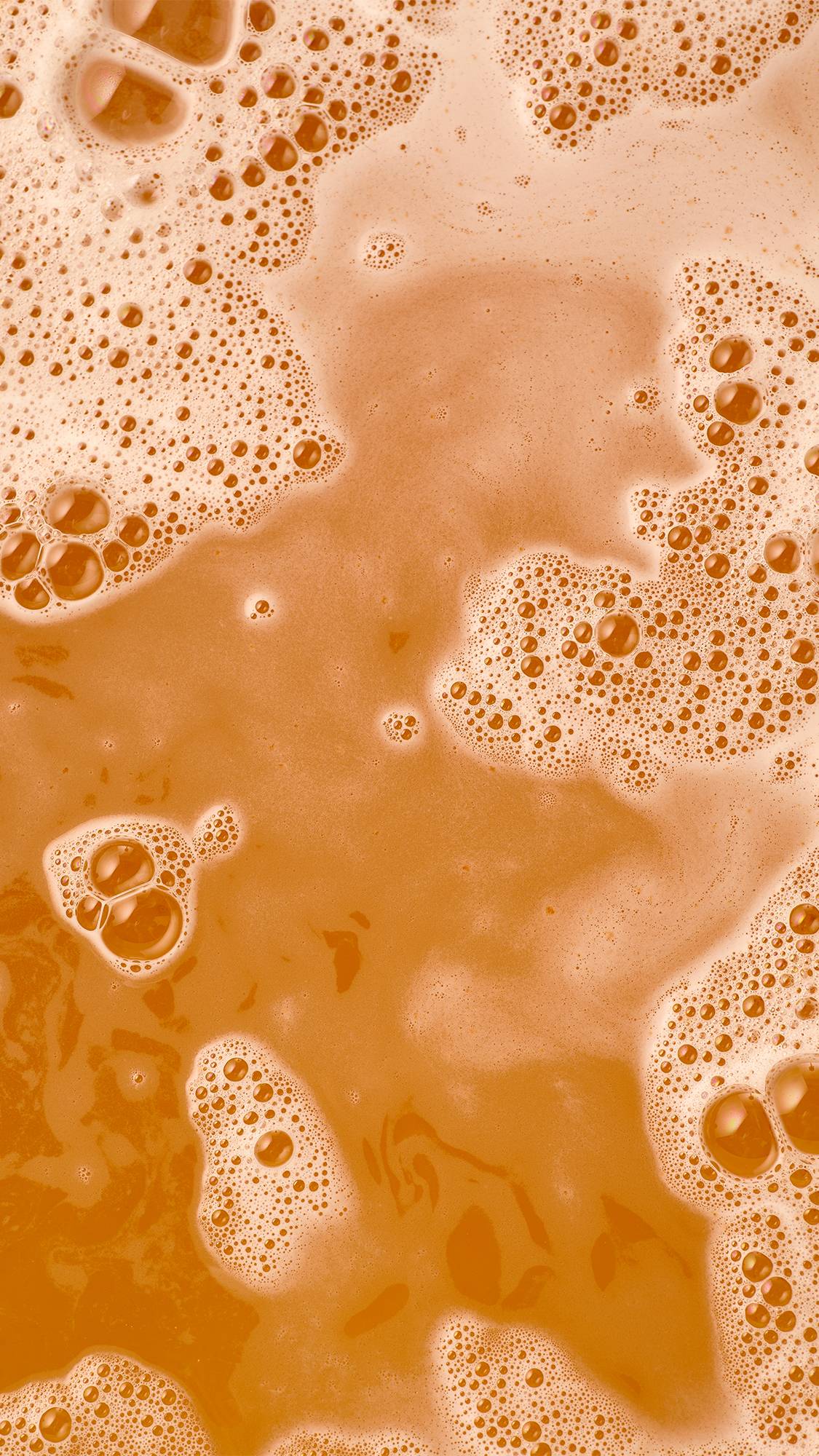 An overhead macro view of warm orange bathwater covered with clusters of white soapy foam, creamy swirls, and delicate bubbles.