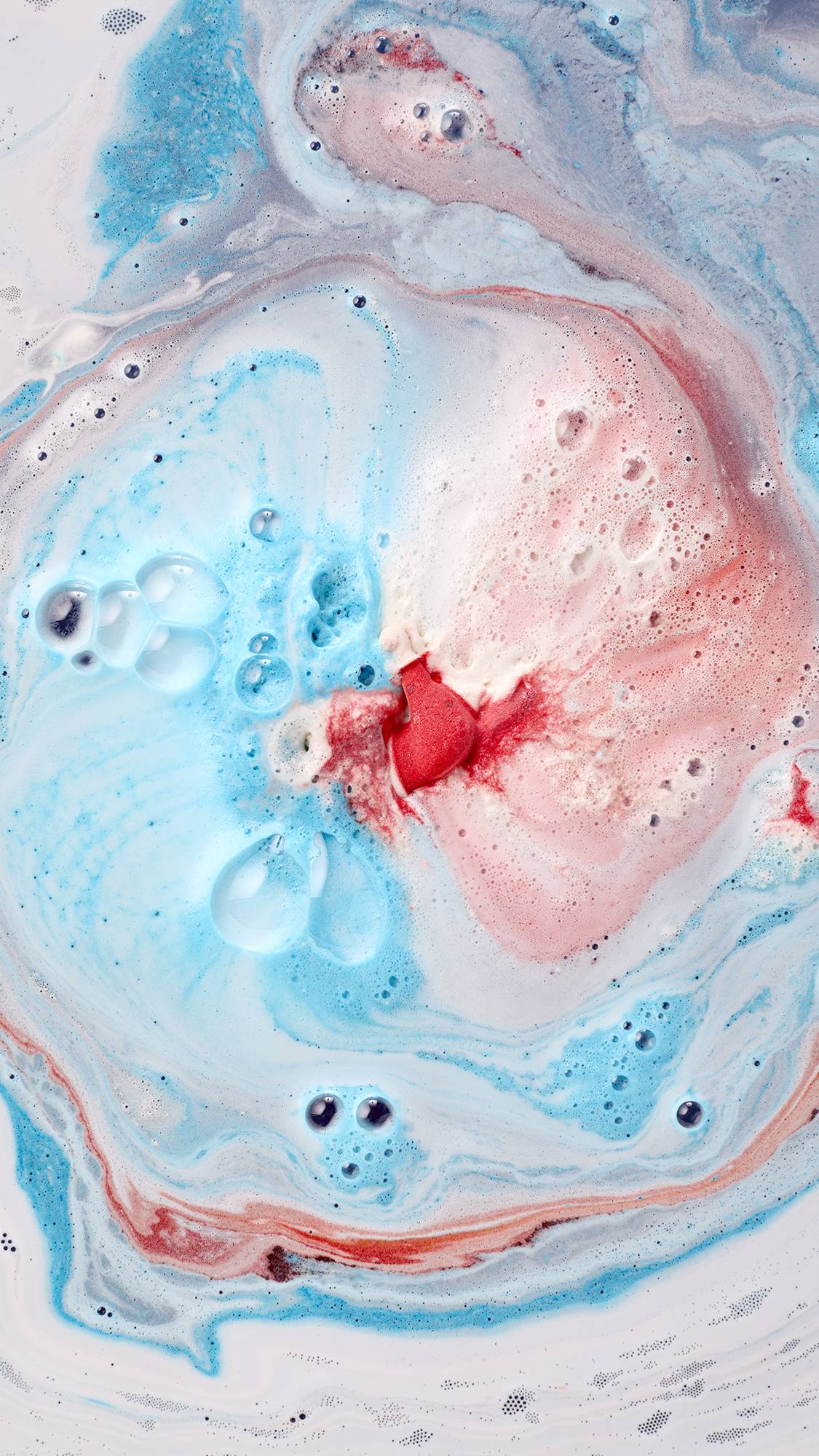 A red fragment of a dissolving Santa bath bomb sits at the centre of swirling light blue and soft pink froth, encircled by dynamic streams of icy blue bathwater and delicate bubbles.