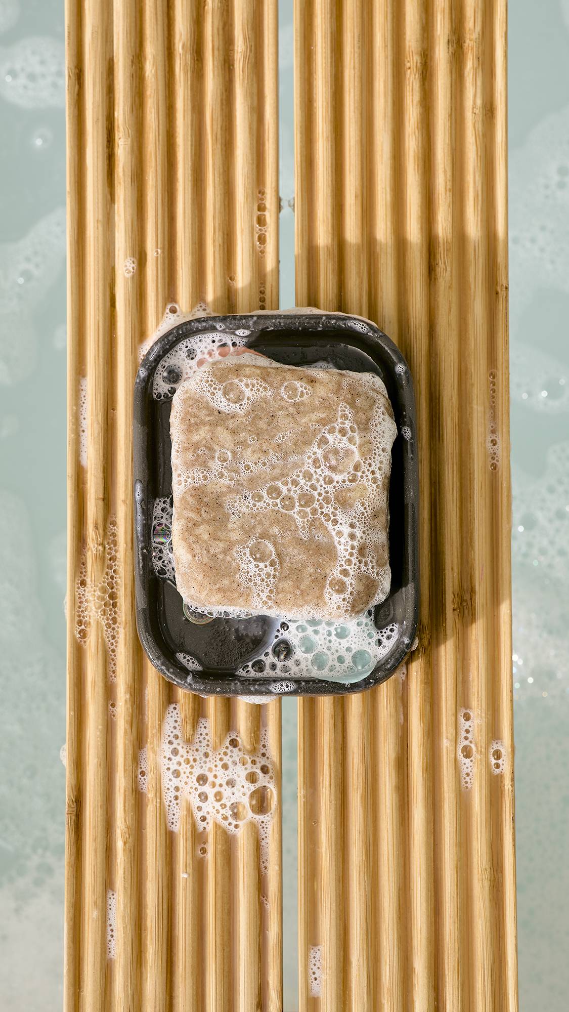 A top-down view of a bar of soapy, textured bar soap in a dark grey ceramic dish with pastel spots, resting on a slatted wooden bath caddy over foamy bathwater.