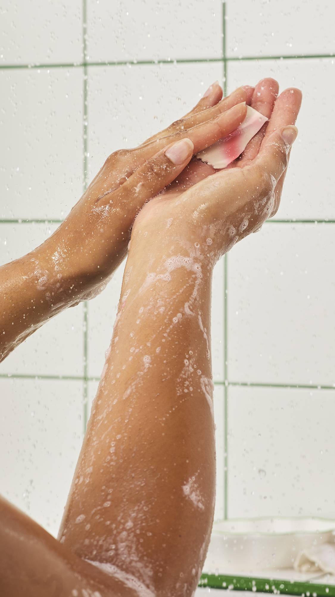 Hands rub together under running shower water, dissolving a small piece of red and white surfactant washing paper to create fine, foamy white bubbles against a white tiled wall background.