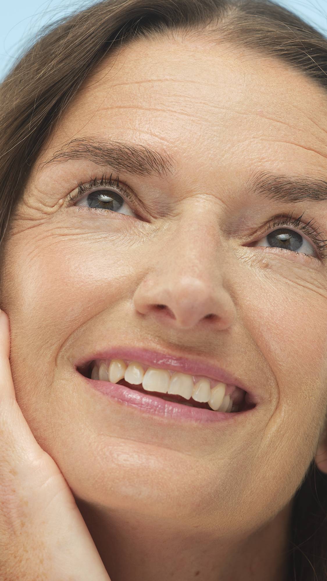 A close-up vertical shot of a smiling person's face, looking up, with a hand resting on their cheek. The Light Medium Warm Beauty Balm provides a smooth, warm base, highlighting their bright eyes.