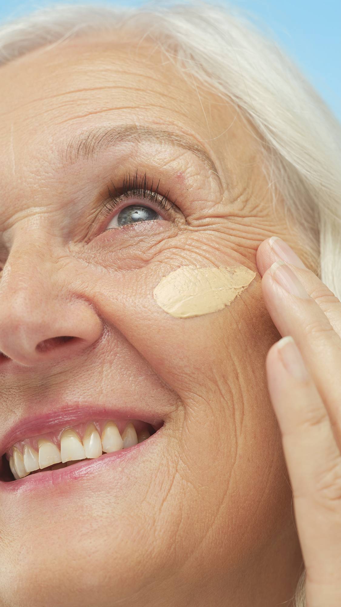 Close-up of a smiling person's cheek as they apply an oval swatch of Light Neutral Skinwear foundation or skin tint with a fingertip.