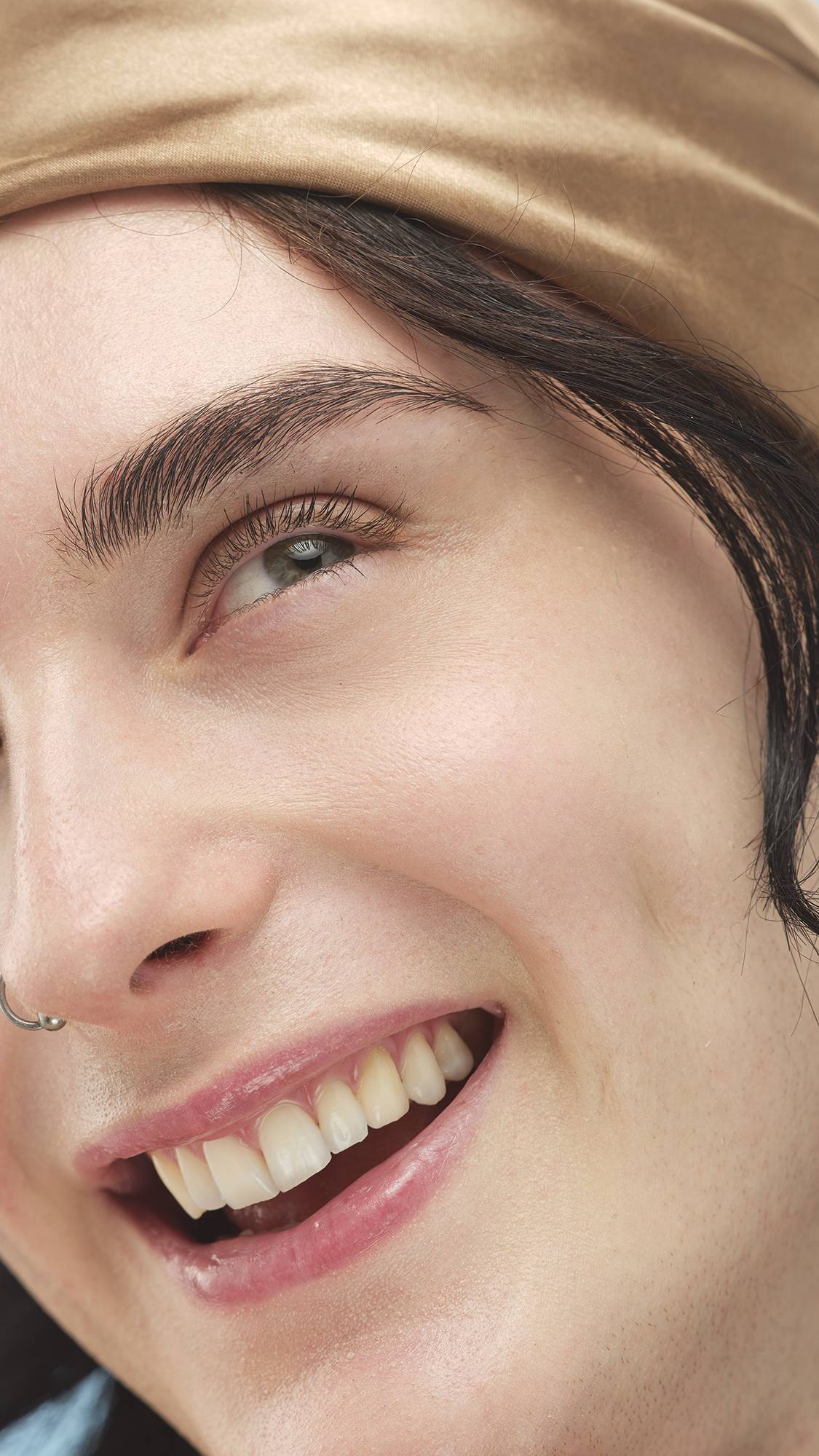 A close-up vertical portrait focusing on a person's smiling face, who is wearing a silky, golden-tan headband and a nose ring. Their skin looks bright and smooth, enhanced by the Light Warm Skinwear.