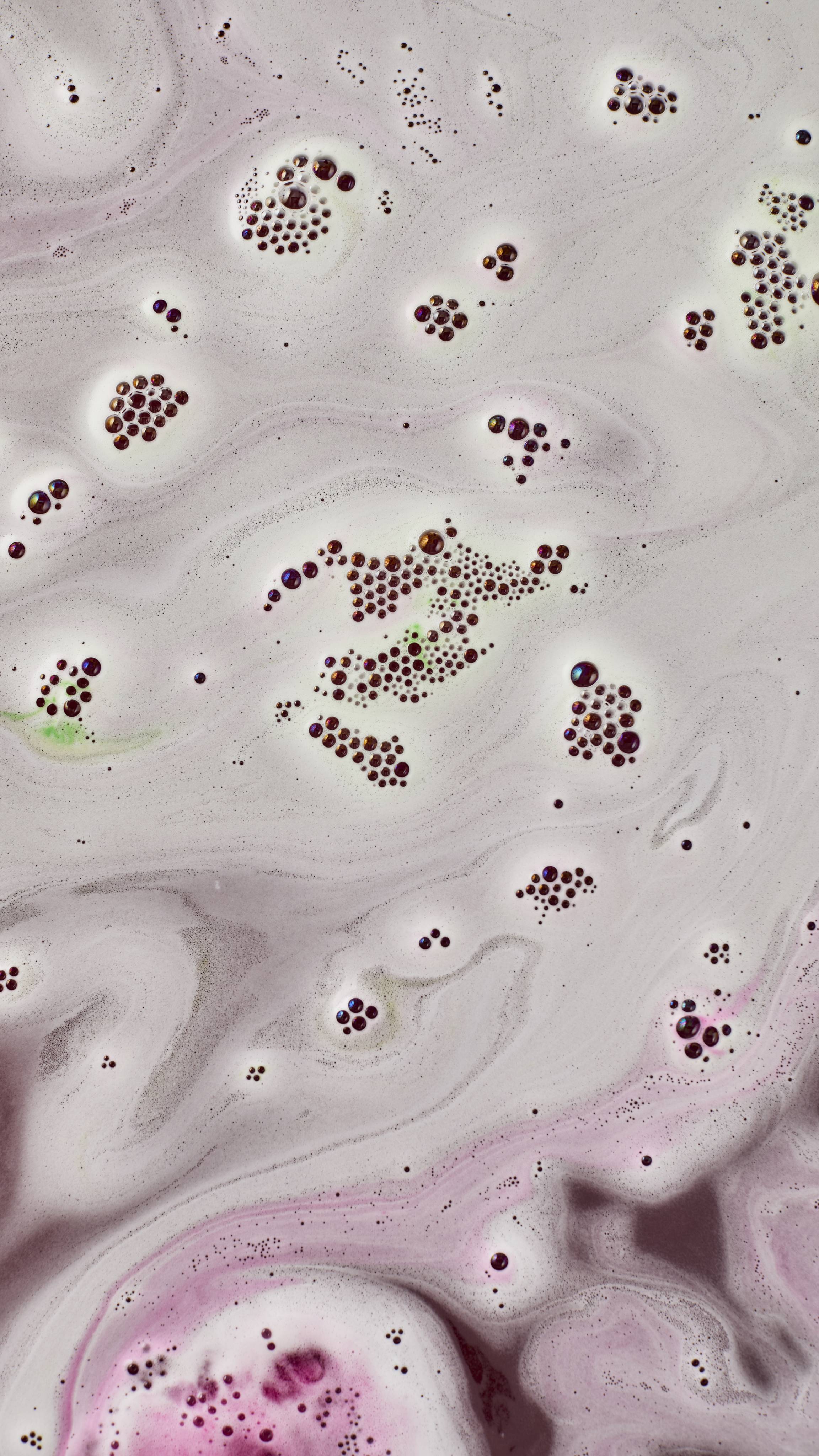 A close-up view of swirled pale lavender and white bath foam, scattered with clusters of dark purple bubbles and subtle hints of lime green and deep pink hue.