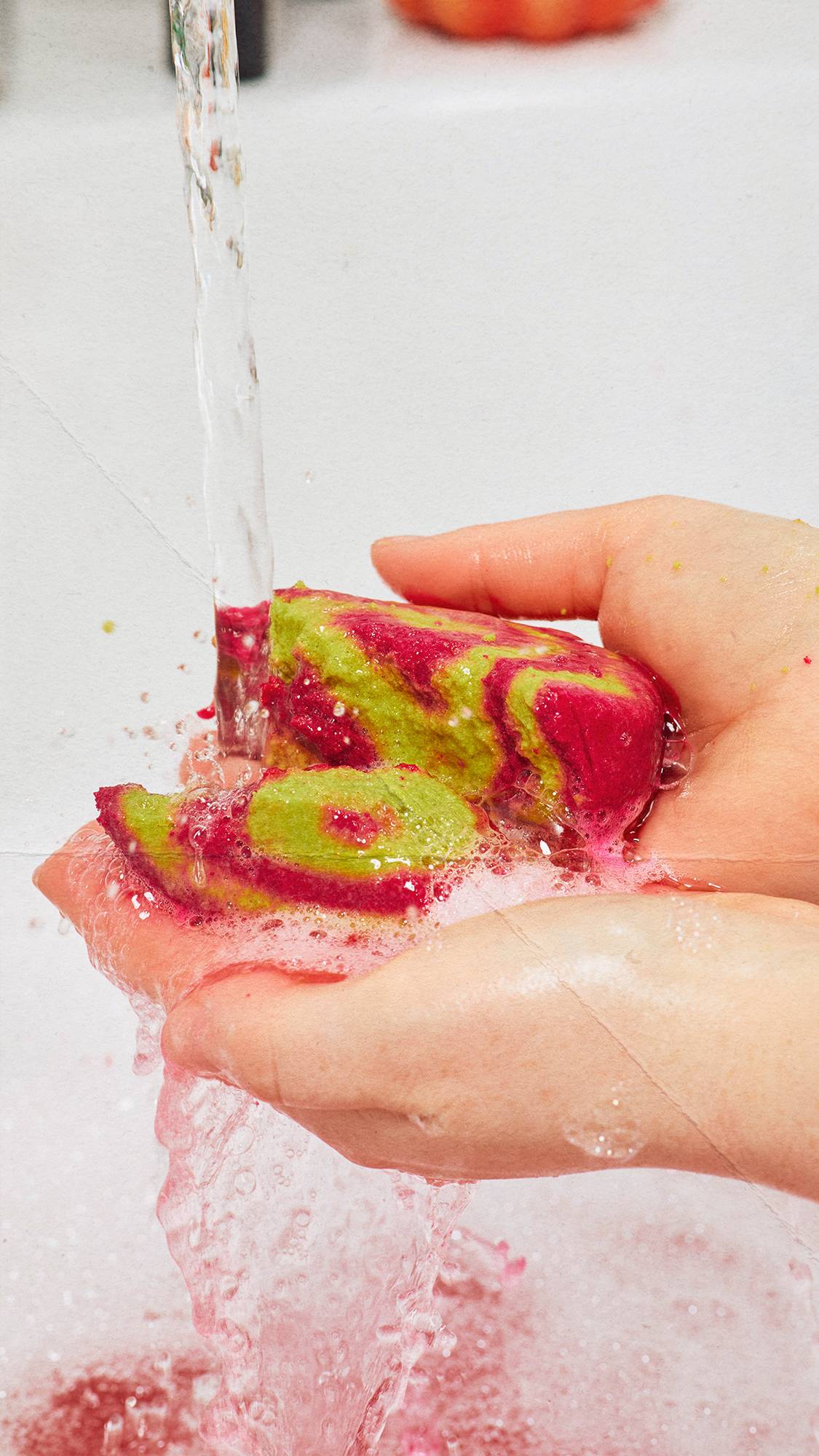 Two hands hold a broken magenta and lime green "Lord of Misrule" bubble bar under a stream of running tap water, creating pink lather and foam as vibrant pink water streams down into a bath.