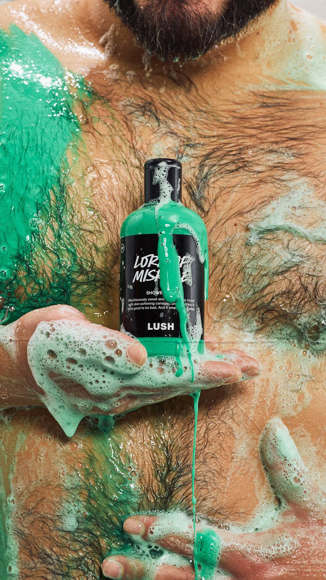 A model with a hairy chest holds a bottle of Lush Lord of Misrule shower gel against their torso, both covered in vivid green shower gel, white suds, and dripping green lather.