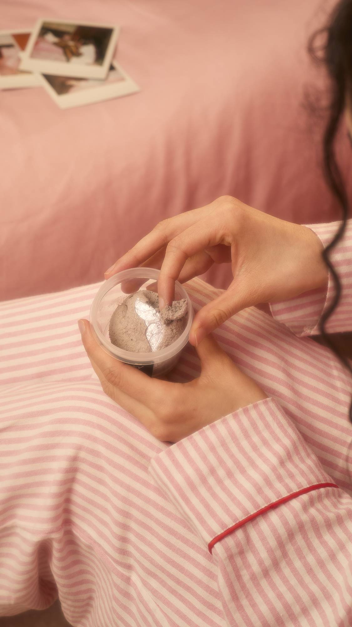 A person wearing pink and white striped pajamas holds a pot of Lovey Dovey facial cleanser, dipping a finger into the product against a soft pink background with polaroid photos.