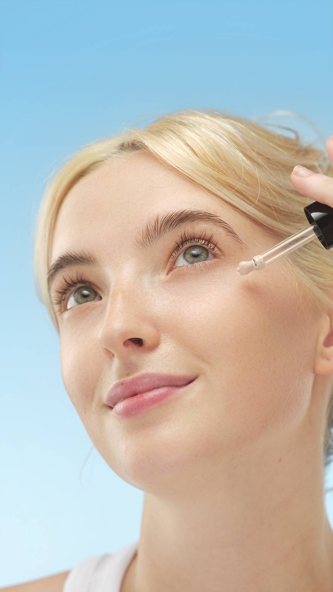 A close-up vertical portrait of a person with fair skin and blonde hair, looking up as they apply a drop of Luminator - skin tint to their cheekbone with a dropper. The skin has a natural sheen, set against a bright blue background.