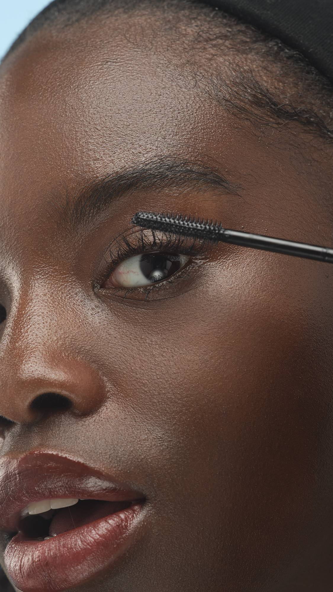 A close-up shot of a person with dark skin applying Lush Lashes ~ Mascara to their upper eyelashes with a black wand. They are wearing a deep, glossy berry-toned lip color and a black headband.