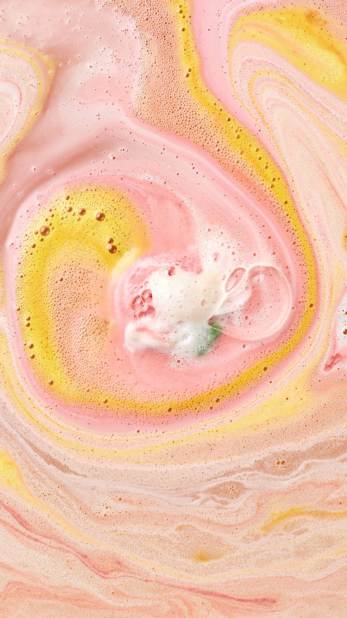 A Christmas pudding bath bomb finishes dissolving in bathwater, creating dynamic swirls of vibrant pink and golden yellow foam around a small central cluster of creamy white froth.
