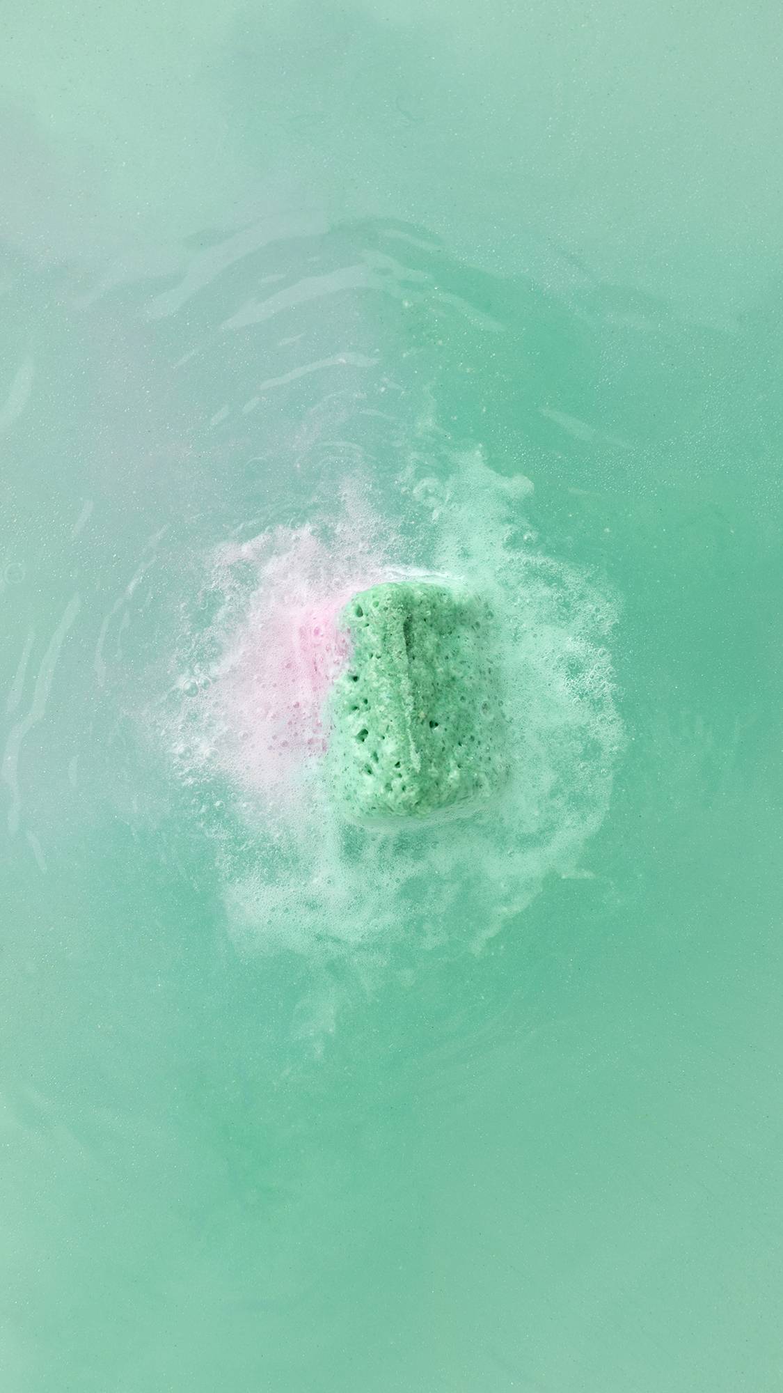 A top-down view of a cube-shaped bath bomb dissolving in mint green water. It creates a vibrant cloud of green and pink foam that swirls into the shimmering, pale green bathwater.