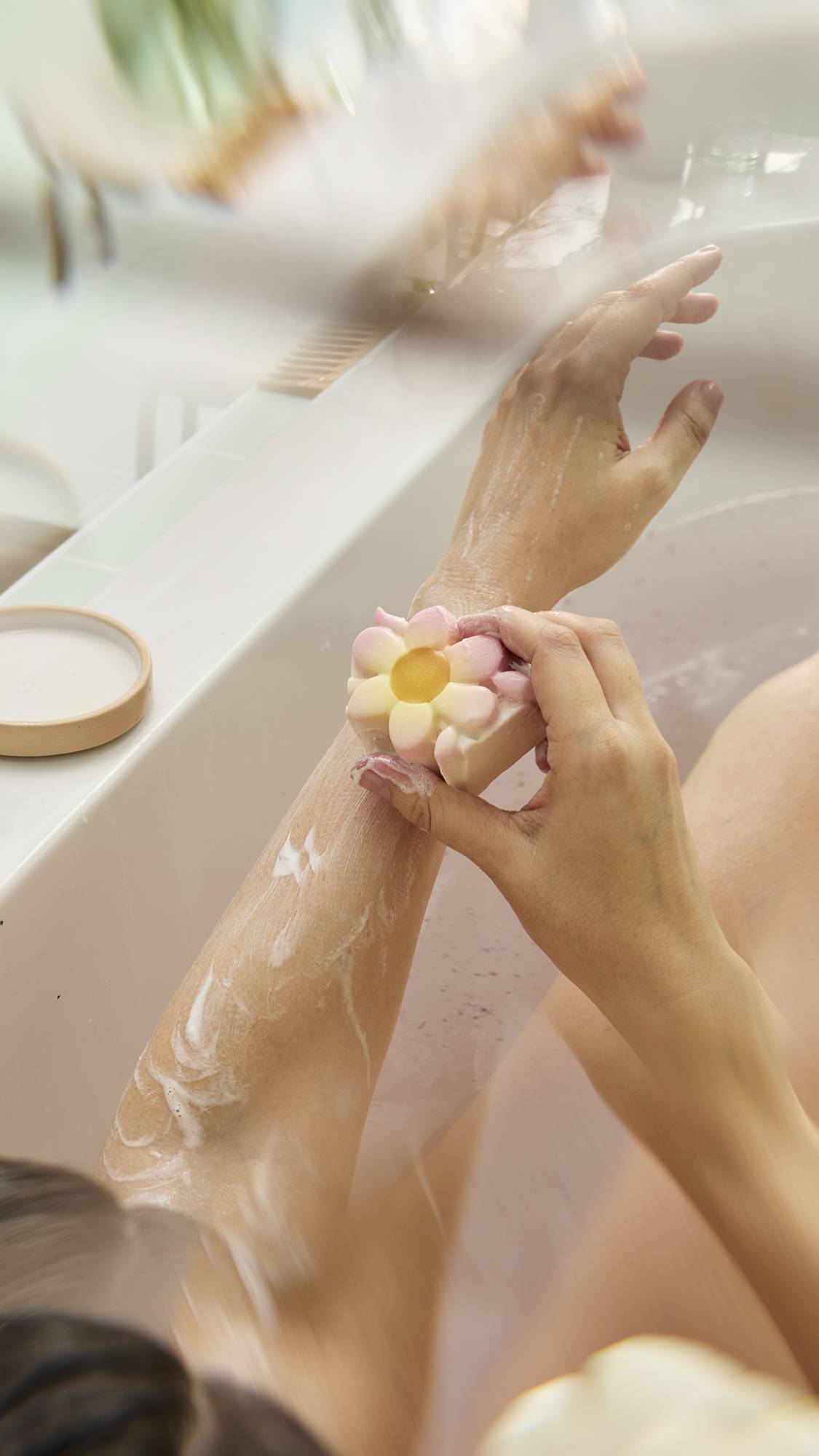 A person massages the flower-shaped "Making Daisy Chains" bar onto their arm, creating a creamy lather. The product features pink-tipped petals and a yellow center, and the scene is set in a bath with a small dish visible on the white ledge.