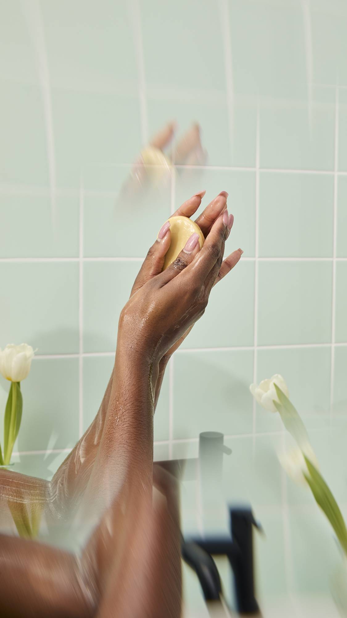 Wet hands hold and massage a "Mama-chino Scrubee" body butter bar under a shower stream against a backdrop of mint green tiles. The scene includes white tulips in the foreground, highlighting the bar being used in a bathing routine.