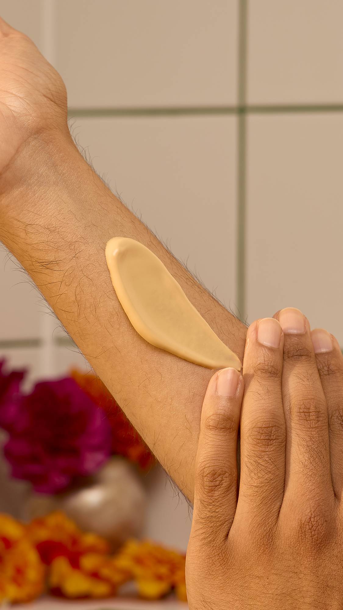 A hand smooths a swatch of rich, yellow Lush "Mango Kulfi" body lotion along an inner forearm against a tiled wall background with blurred flowers.