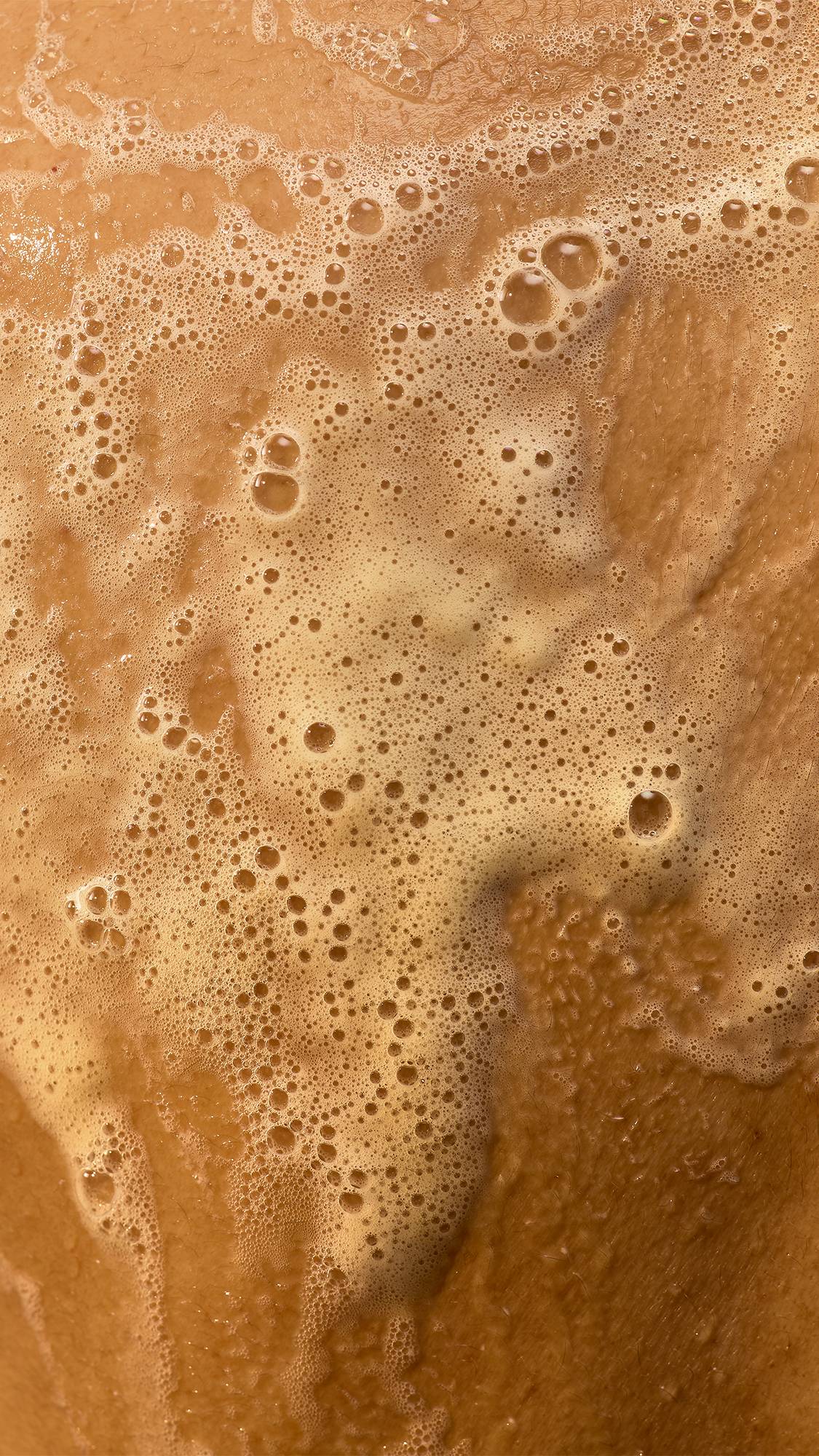 A close-up shot of rich, creamy golden "Mango Lassi" shower gel lather with fine bubbles smooths across wet skin, creating a warm, foamy texture.