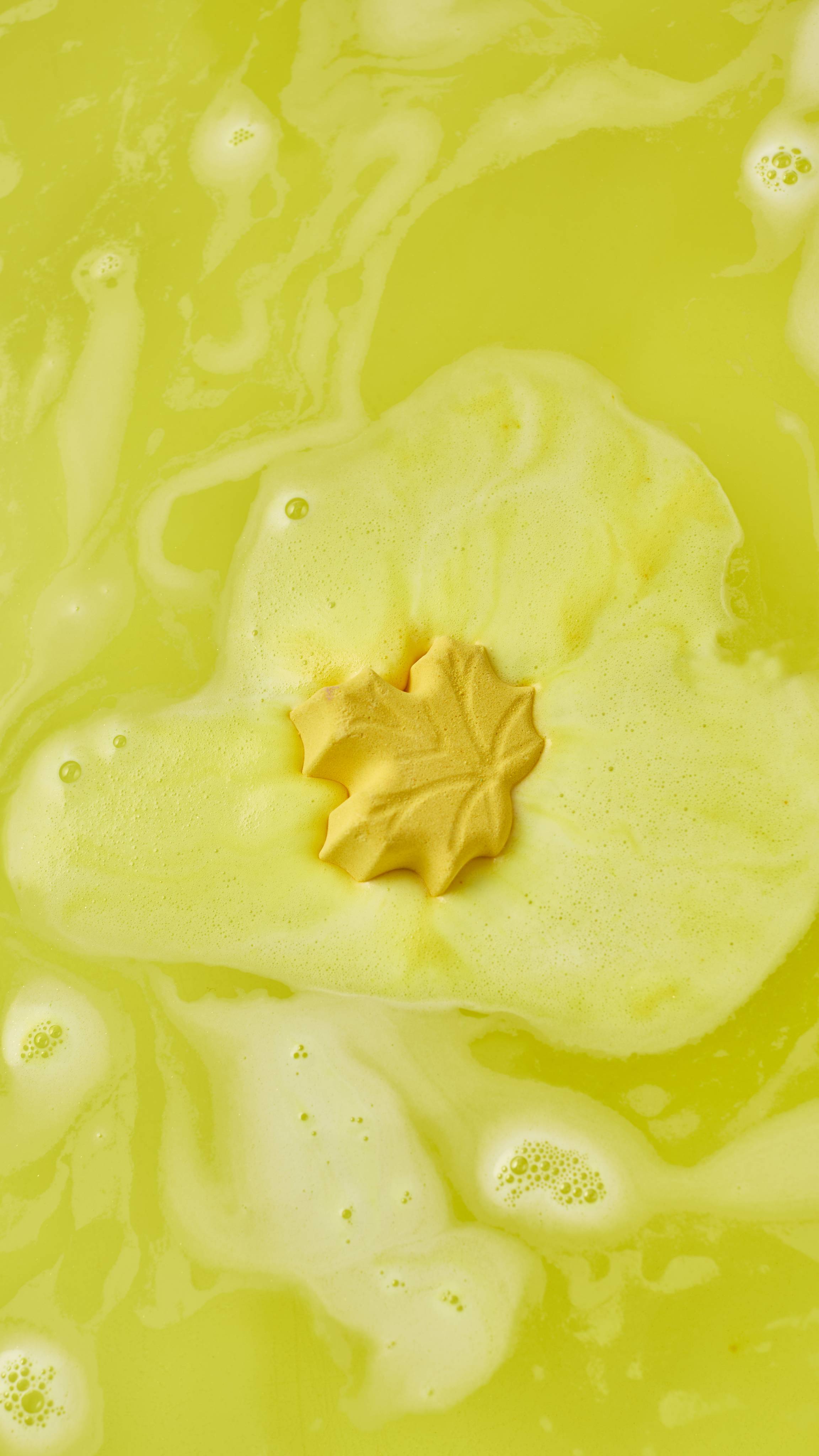A top-down view of a small yellow leaf-shaped bath bomb dissolving in the middle of a pool of creamy yellow foam, surrounded by bright, sunny-yellow swirling bathwater.