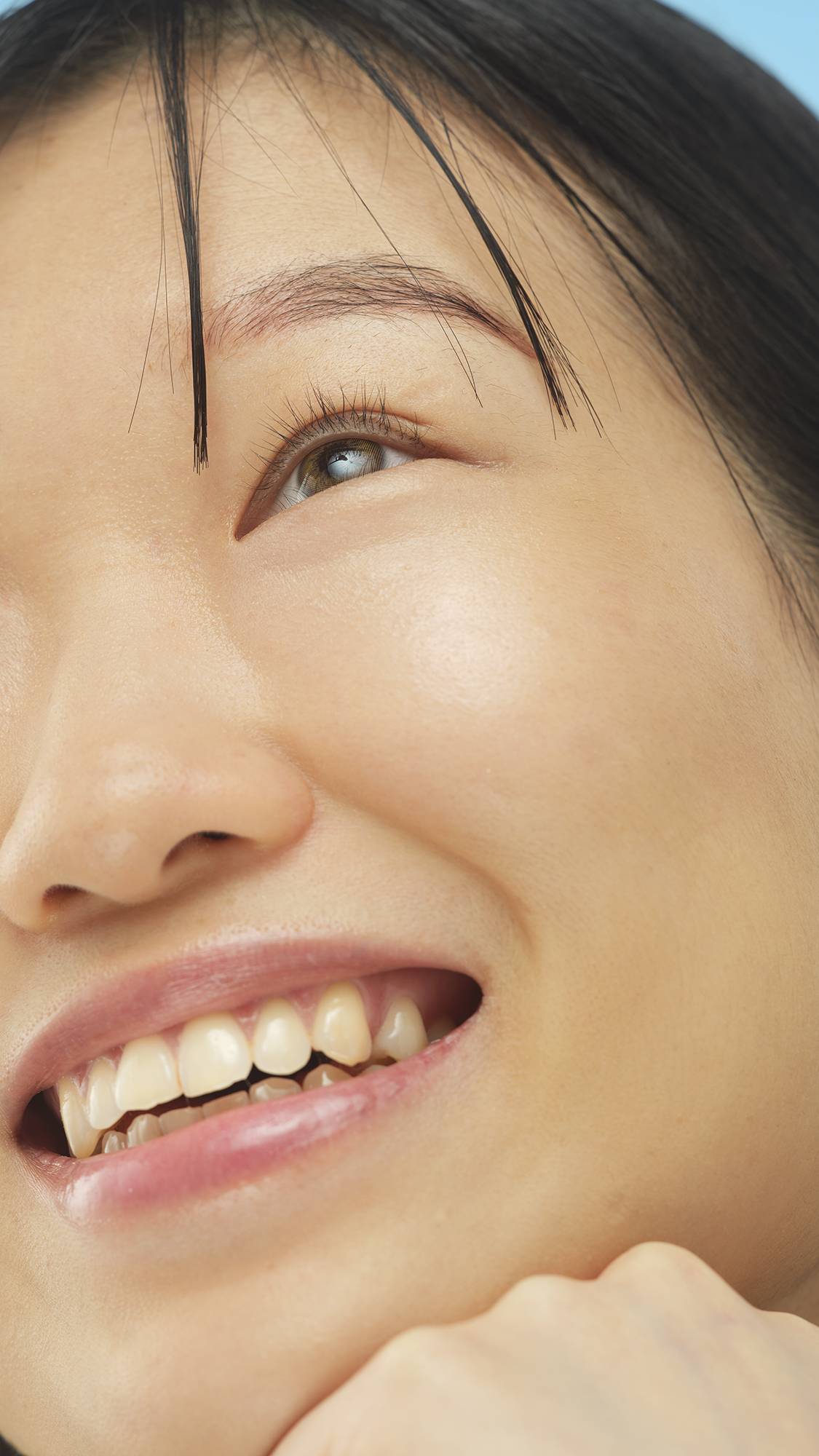 Close-up on a person's smiling face, showing a dewy complexion from the Medium Cool Skinwear foundation. The person looks upward, with their chin resting on their hand.
