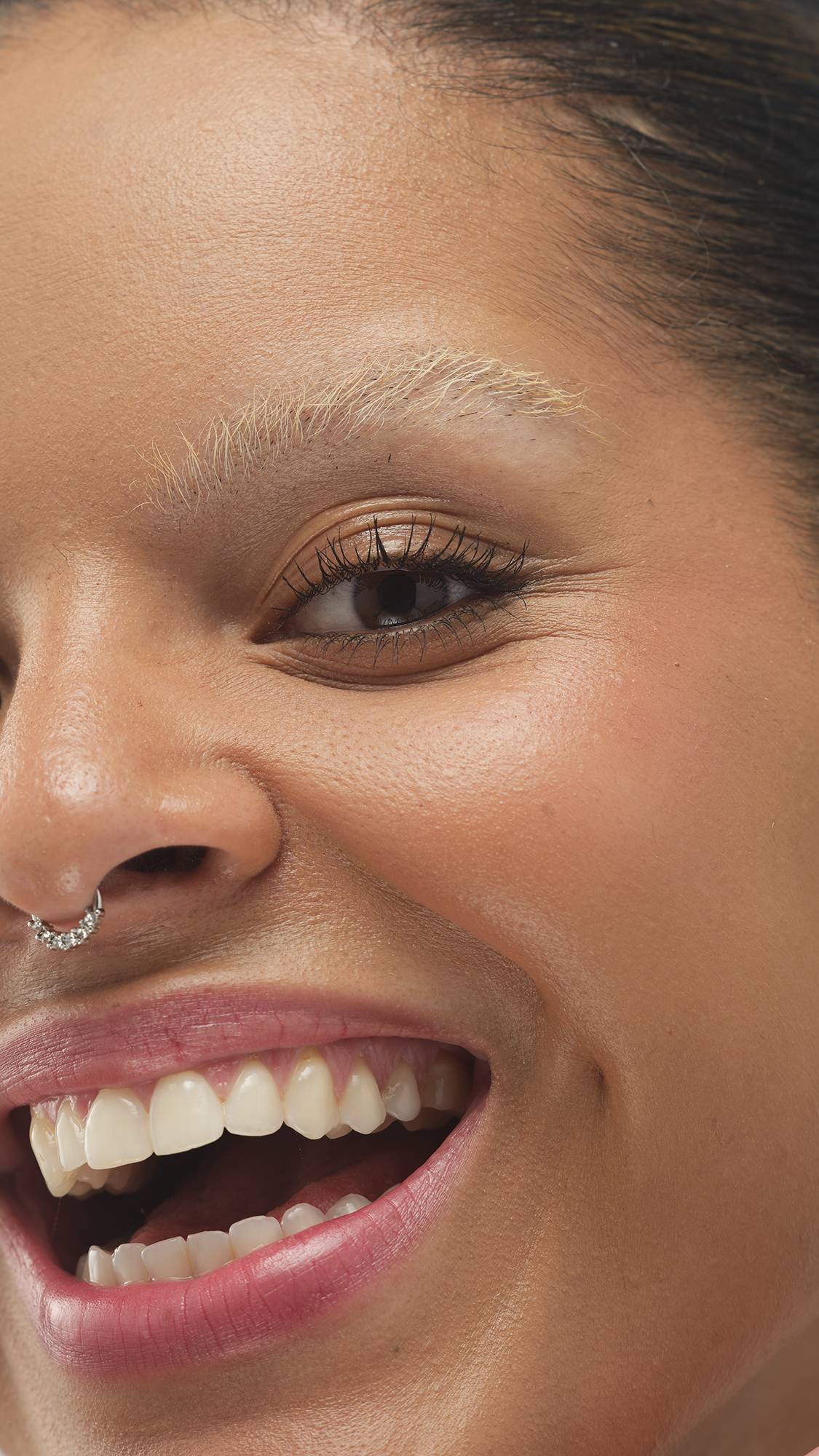 A close-up vertical shot of a person's smiling face, showing their eye, cheek, and open mouth, highlighting their bleached-blonde eyebrow and a septum piercing. The Medium Dark Cool Beauty Balm provides a subtle, natural glow.



