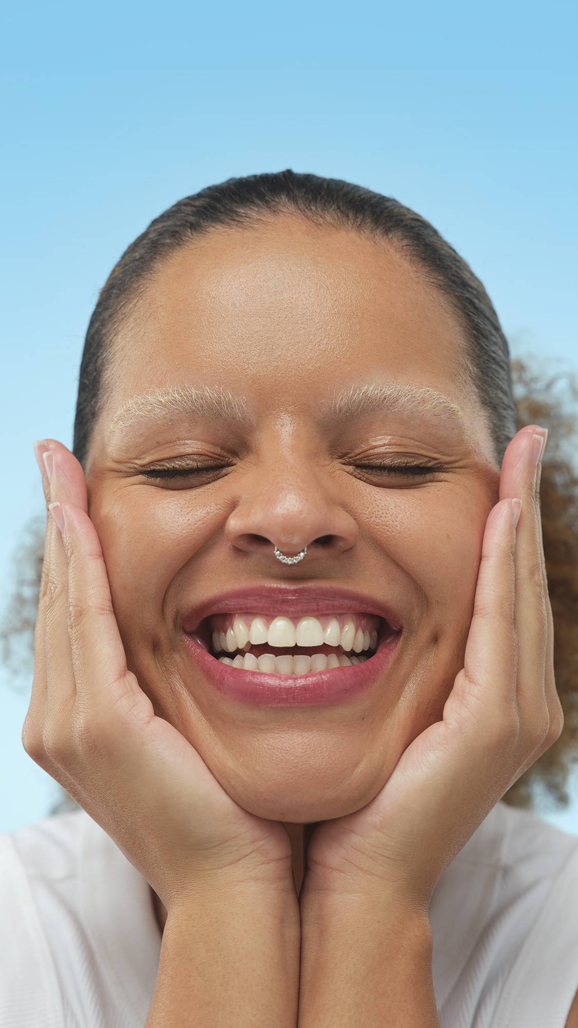 A person with bleached eyebrows applies a Medium Dark Cool Skinwear foundation or skin tint to their cheek with a finger, posing with a wide smile against a light blue background.