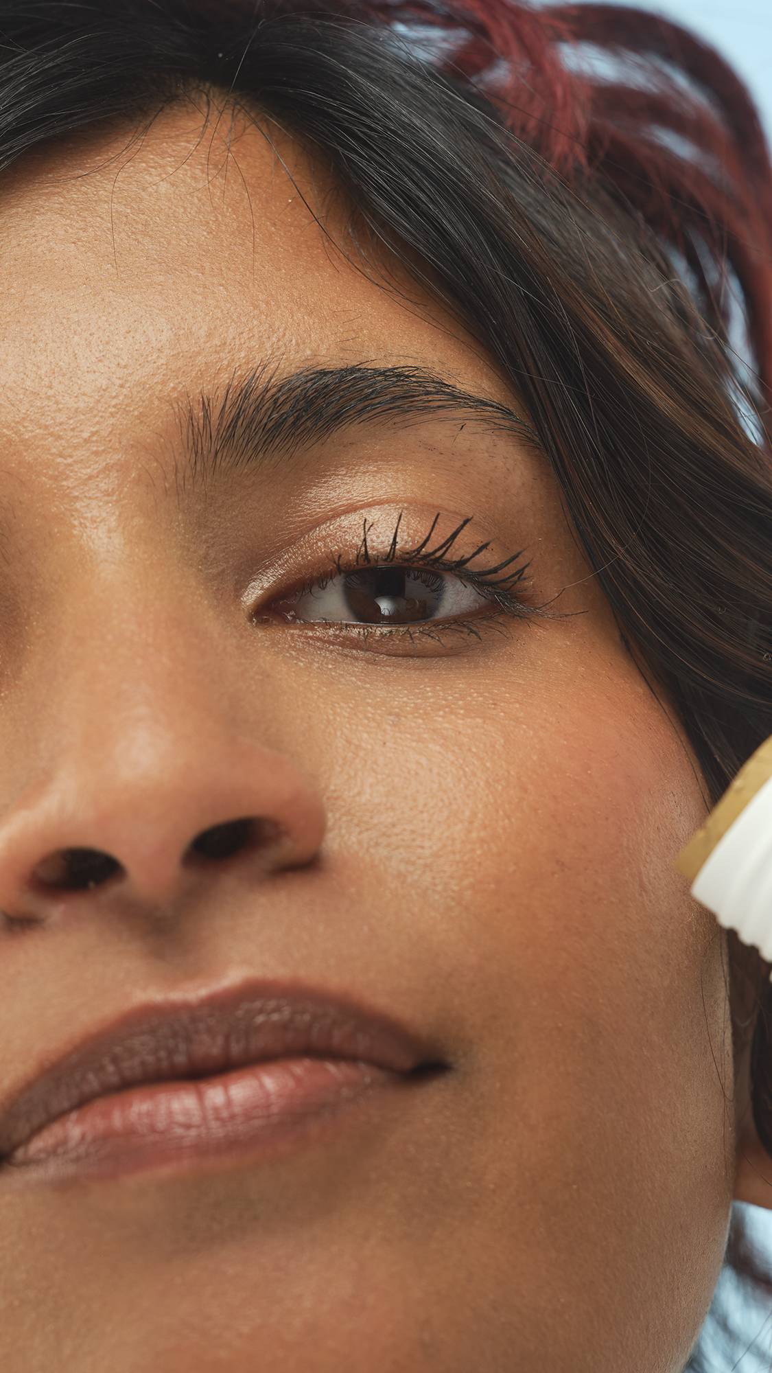 A close-up vertical shot of a person's eye, cheek, and lips. The medium-dark skin has a subtle glow from the Medium Dark Neutral Beauty Balm, and part of the white product packaging is visible near the cheek.