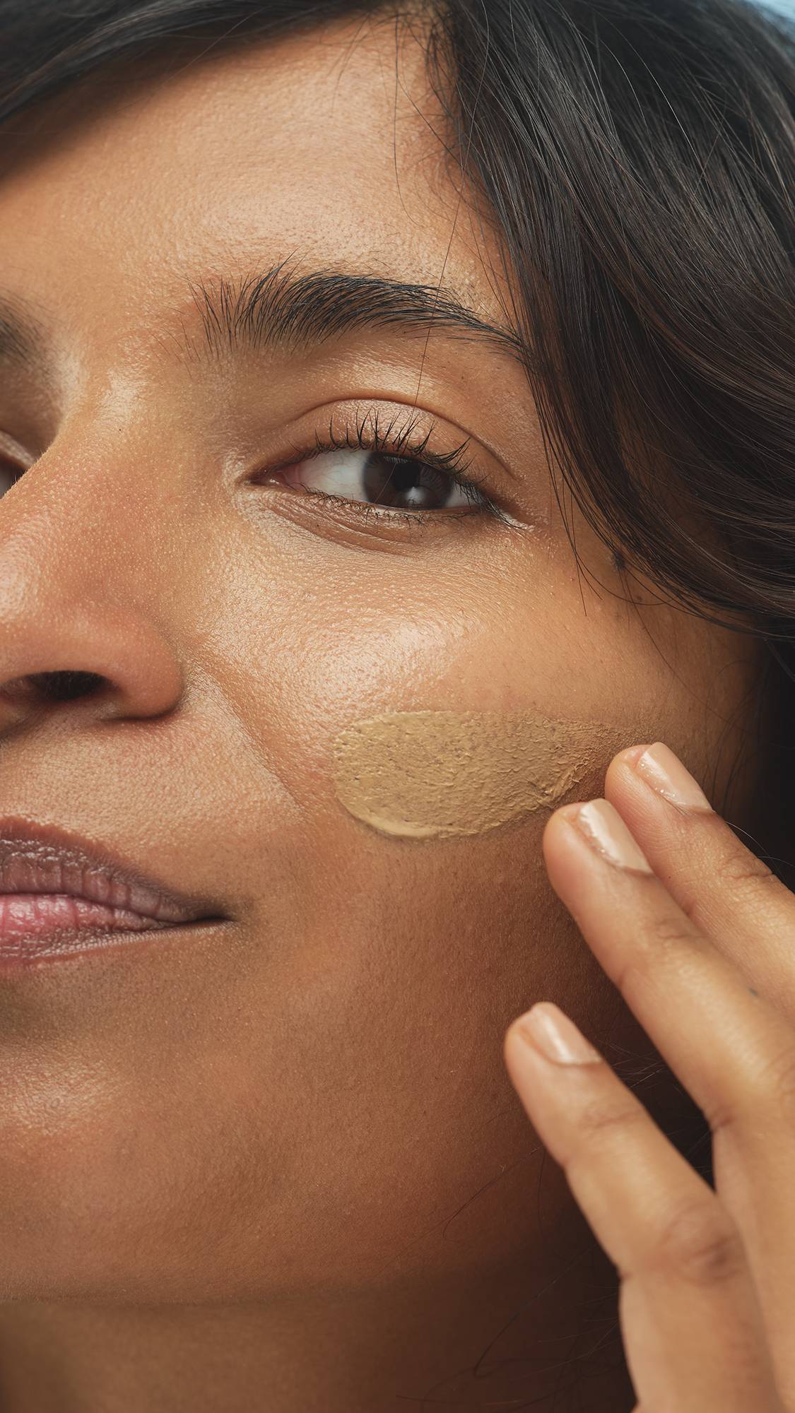 Close-up of a person's cheek as they apply a Medium Dark Neutral Skinwear foundation or skin tint with their finger in a smooth, thick oval swatch.