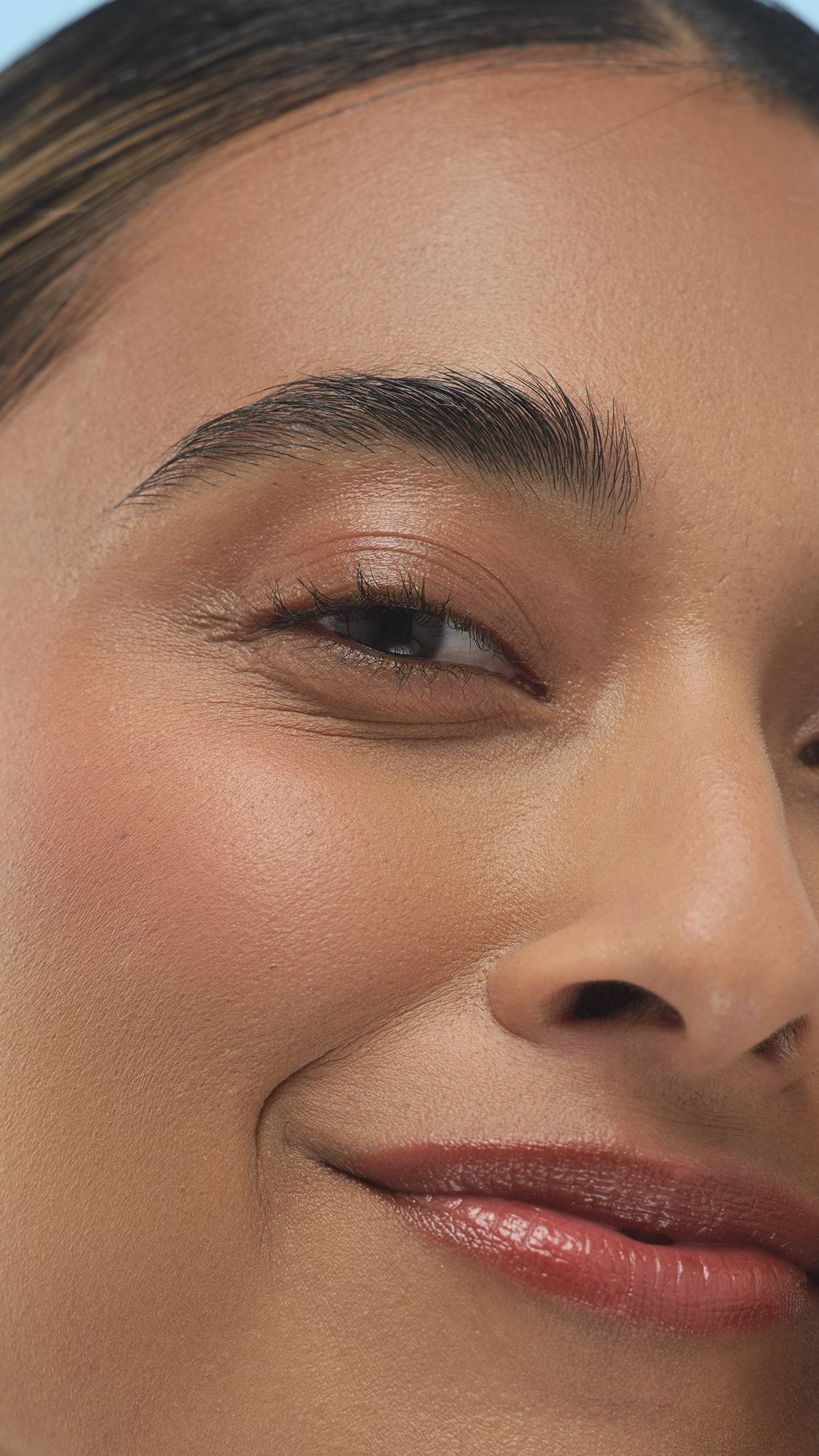 A close-up vertical shot of a person's smiling upper face, showing a subtle flush and glow from the Medium Dark Warm Beauty Balm on their cheeks. They have smooth, warm-toned skin and well-groomed brows.