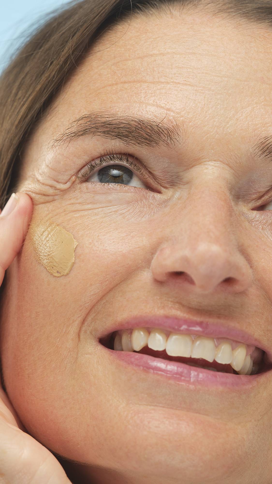 Close-up on a smiling person's cheek as they apply a smooth, circular swatch of Medium Neutral Skinwear foundation or skin tint with a finger.