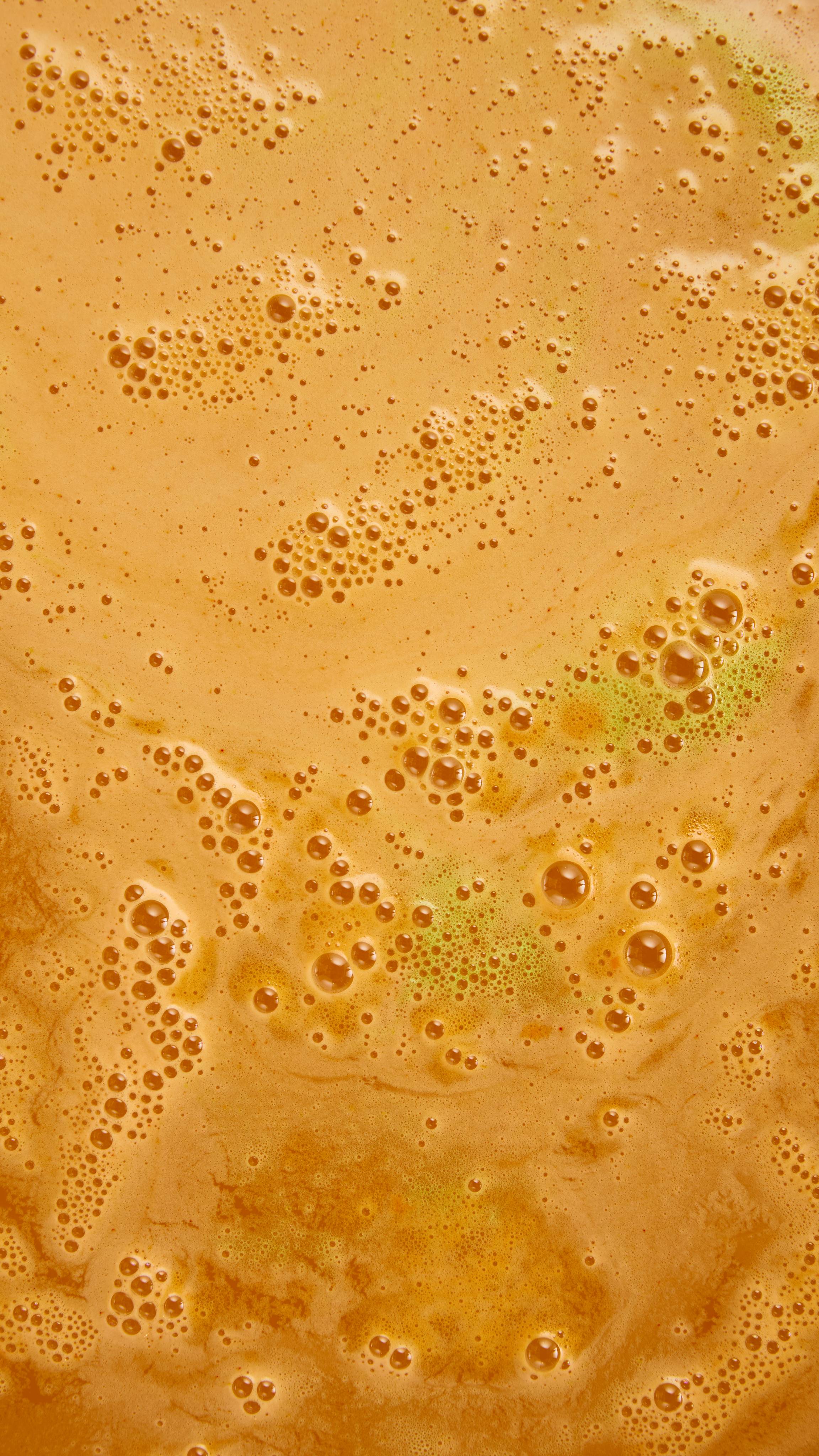 A close-up view of vibrant orange bathwater covered in dense clusters of fine bubbles with subtle lime-green swirls.