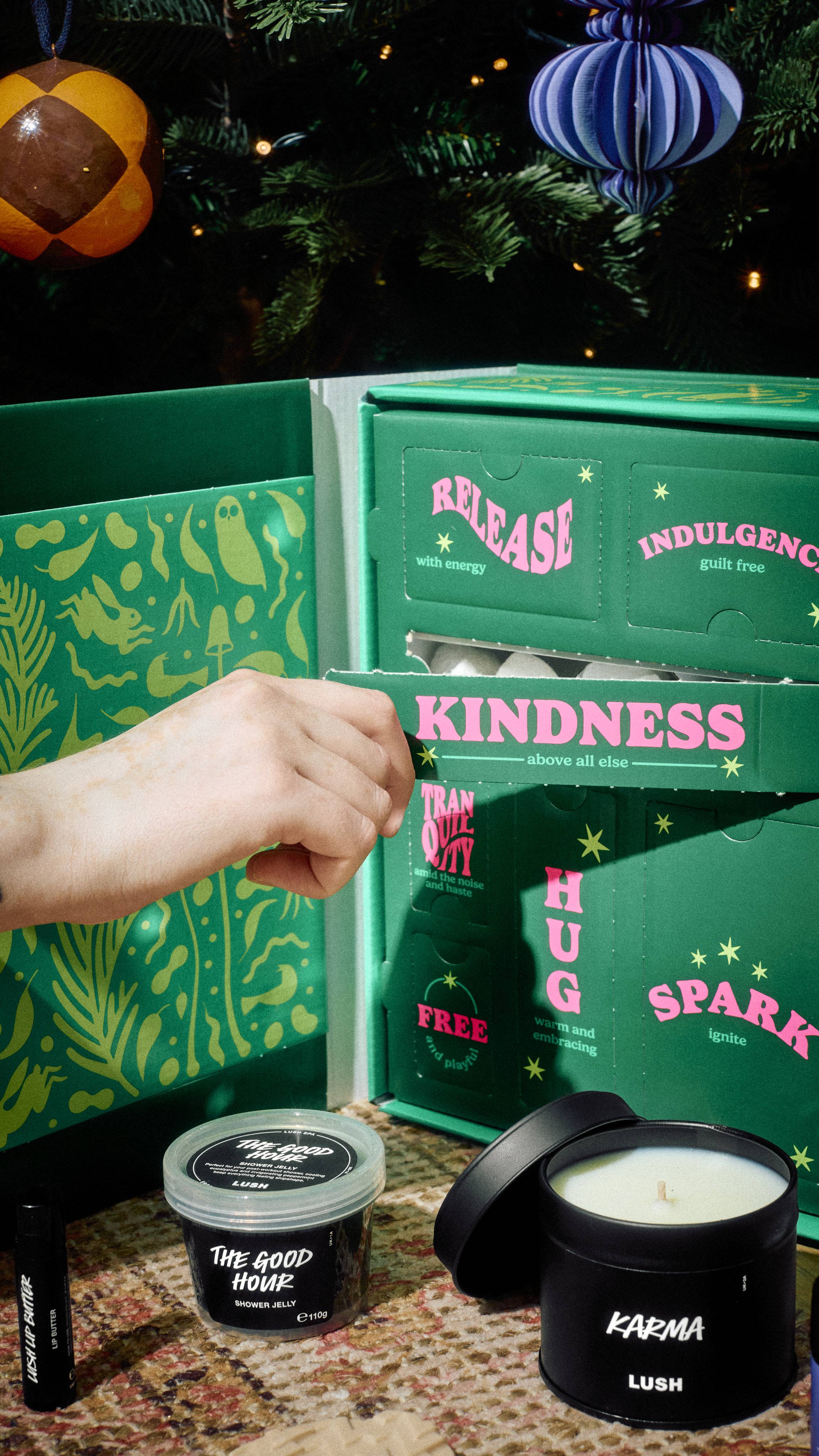 A hand opens a compartment labelled Kindness on the green Lush Mindful Moments gift cabinet, with a Karma candle, lip butter, and The Good Hour shower jelly set out on a patterned rug below a decorated Christmas tree.