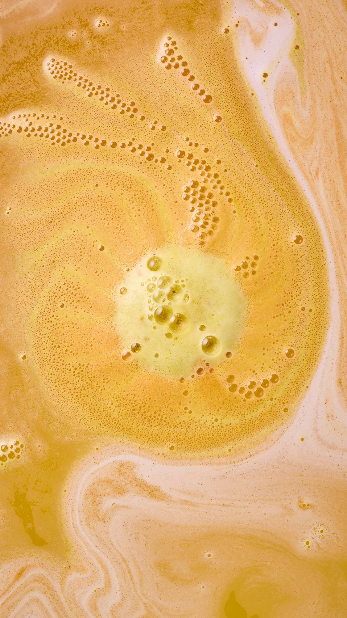 An overhead close-up of a "Mooncake" bath bomb fully dissolving into water. A bright yellow pool of thick, bubbly foam sits at the centre of a swirling vortex of warm orange and creamy peach water, forming distinct paths of tiny bubbles.