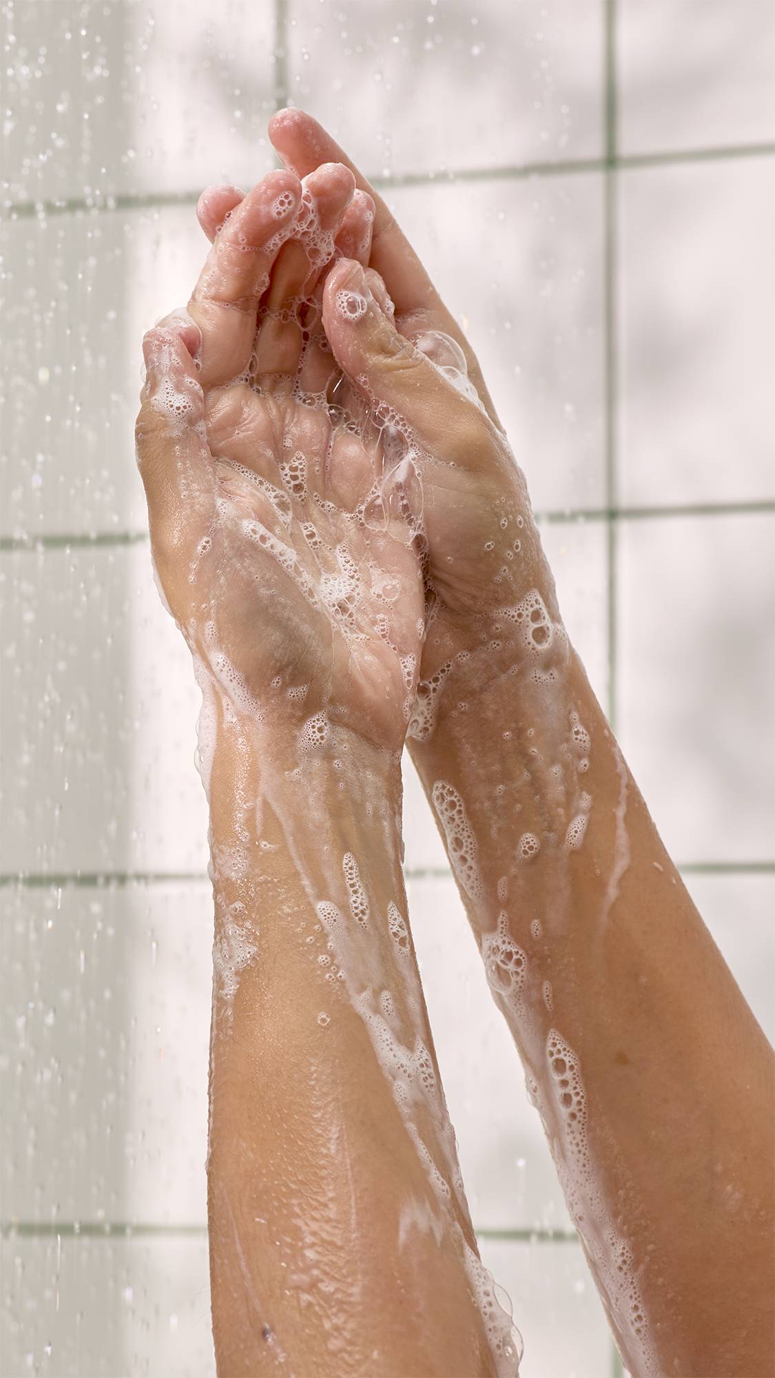 Two soapy hands and arms covered in rich white foam lathering together under cascading water in a shower against a white tiled wall.   