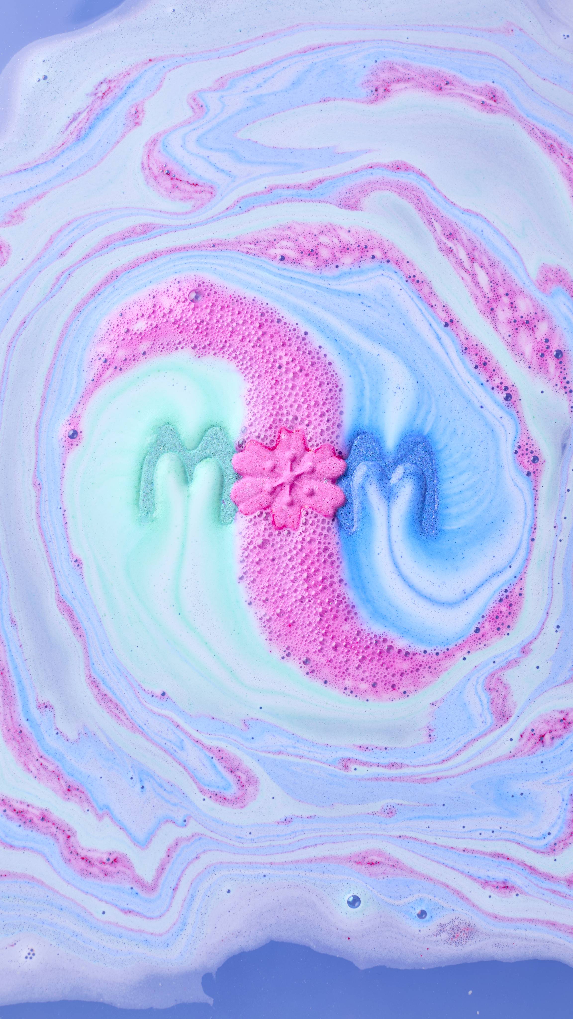 An aerial view image shows a blue, green, and pink "MUM" bath bomb. The coloured letters create a vibrant, swirling spiral of pink, blue, and mint green foam that marbles together across the surface of the pale blue bathwater