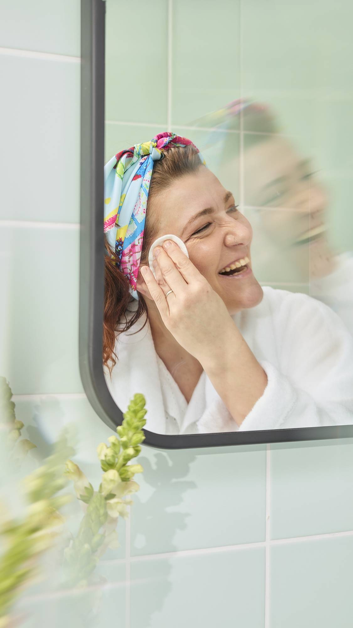 The image shows a model with wavy auburn hair in a bathroom mirror, smiling while cleansing her face with a cotton pad. She has fashioned the colourful "My Family Houses" fabric knot wrap into a stylish headband.